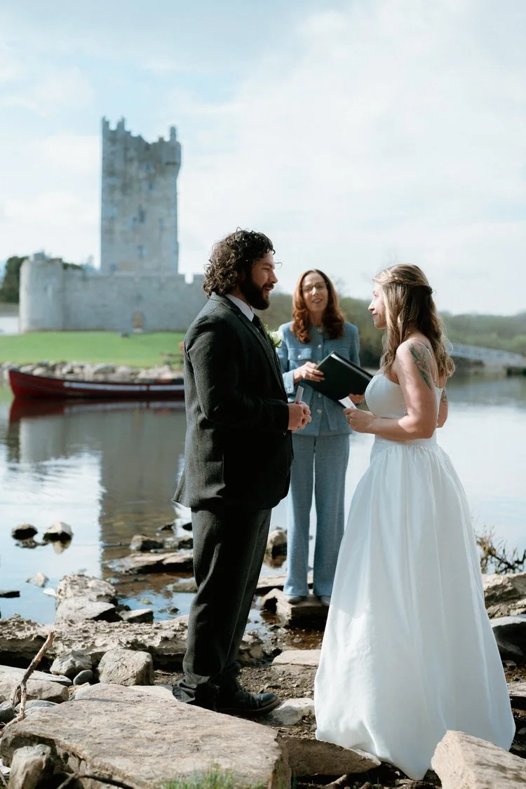A couple getting married outdoors by a river with a castle in the background, standing on rocks and exchanging vows with a woman officiant - Irish wedding planner ceremony