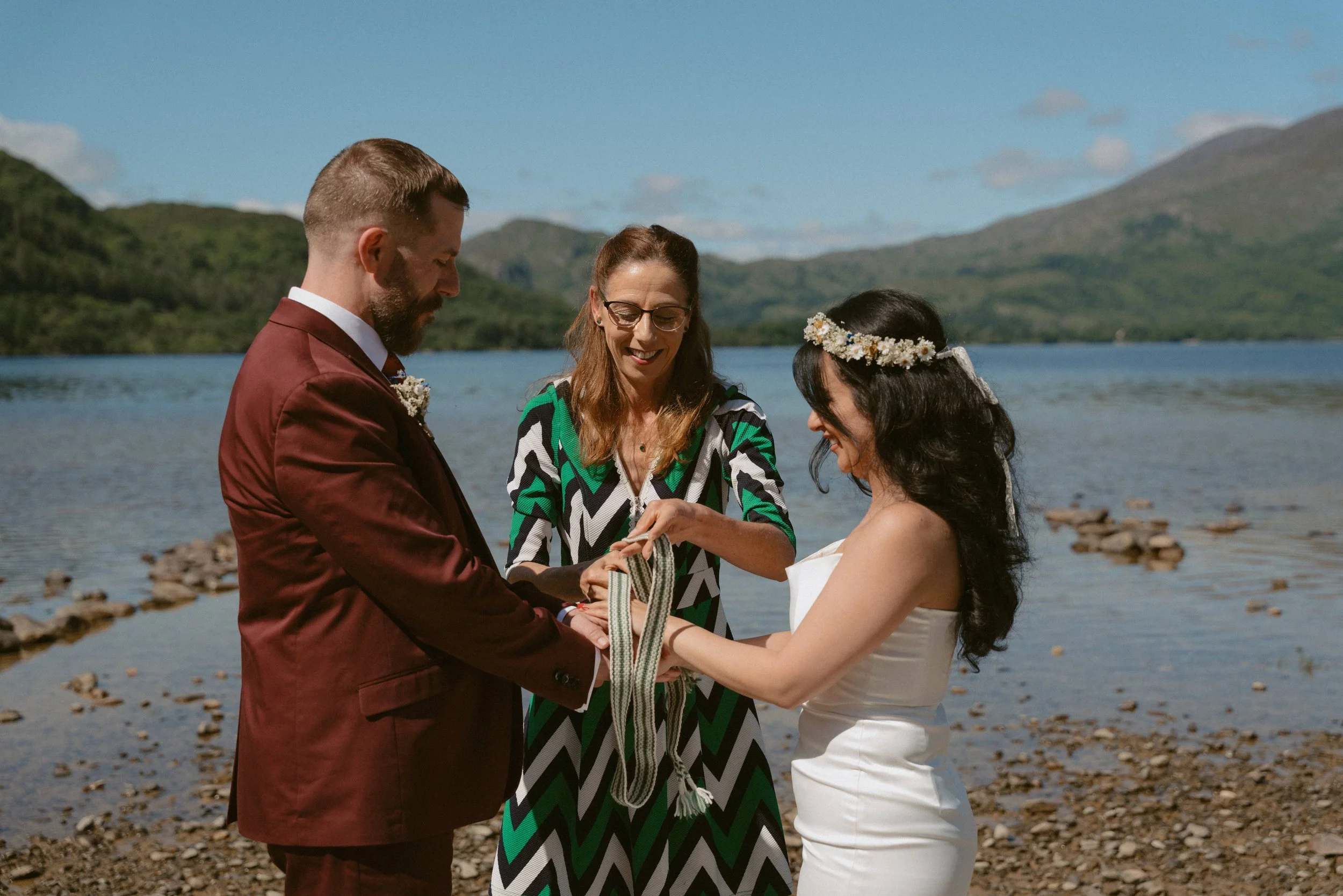 A wedding ceremony taking place outdoors by a lake, with a couple exchanging rings and a woman officiant present. The background features mountains and a partly cloudy blue sky - destination wedding in Ireland
