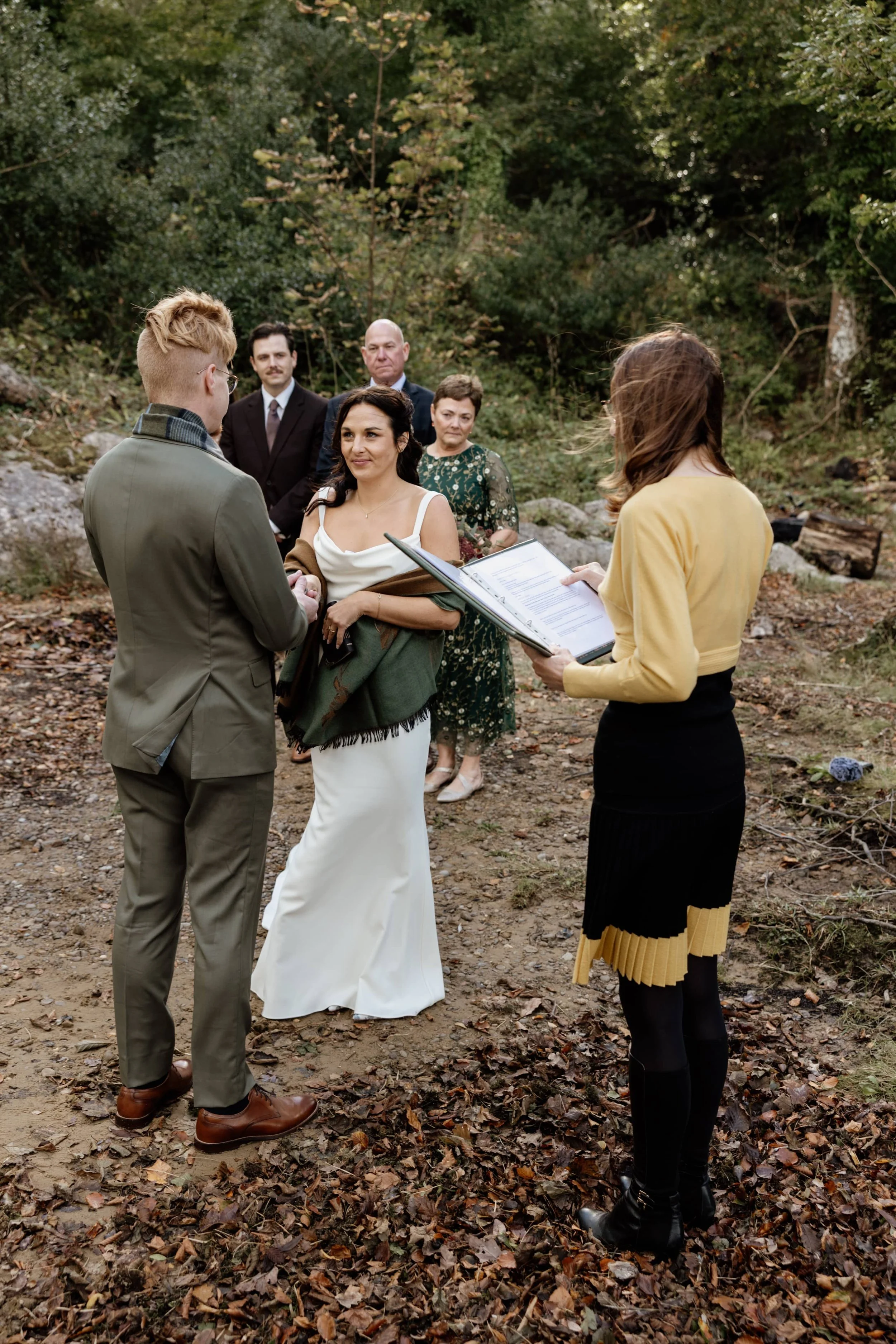 A couple eloping in Ireland in an outdoor wooded setting with the bride and groom exchanging vows, surrounded by officiants and guests