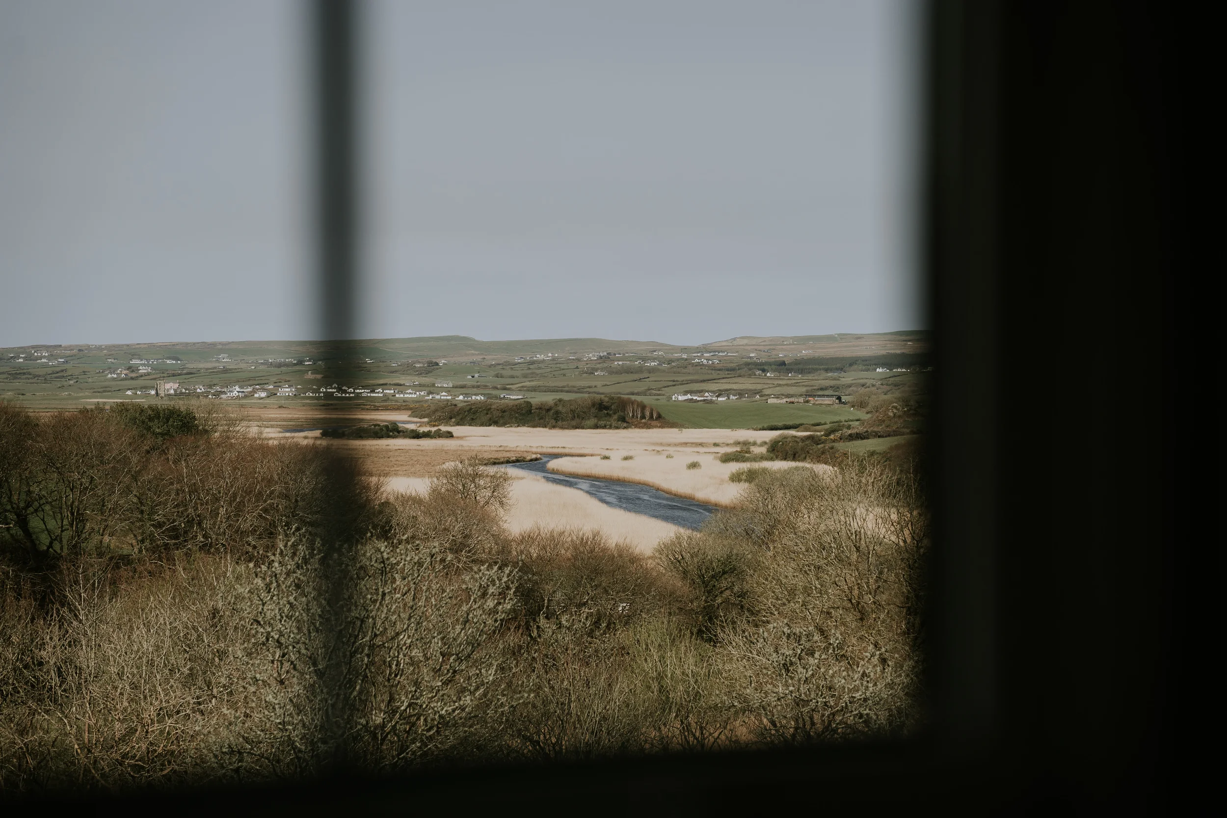 A scenic view of a river winding through fields and trees, with a distant village on rolling hills, seen through a window frame.