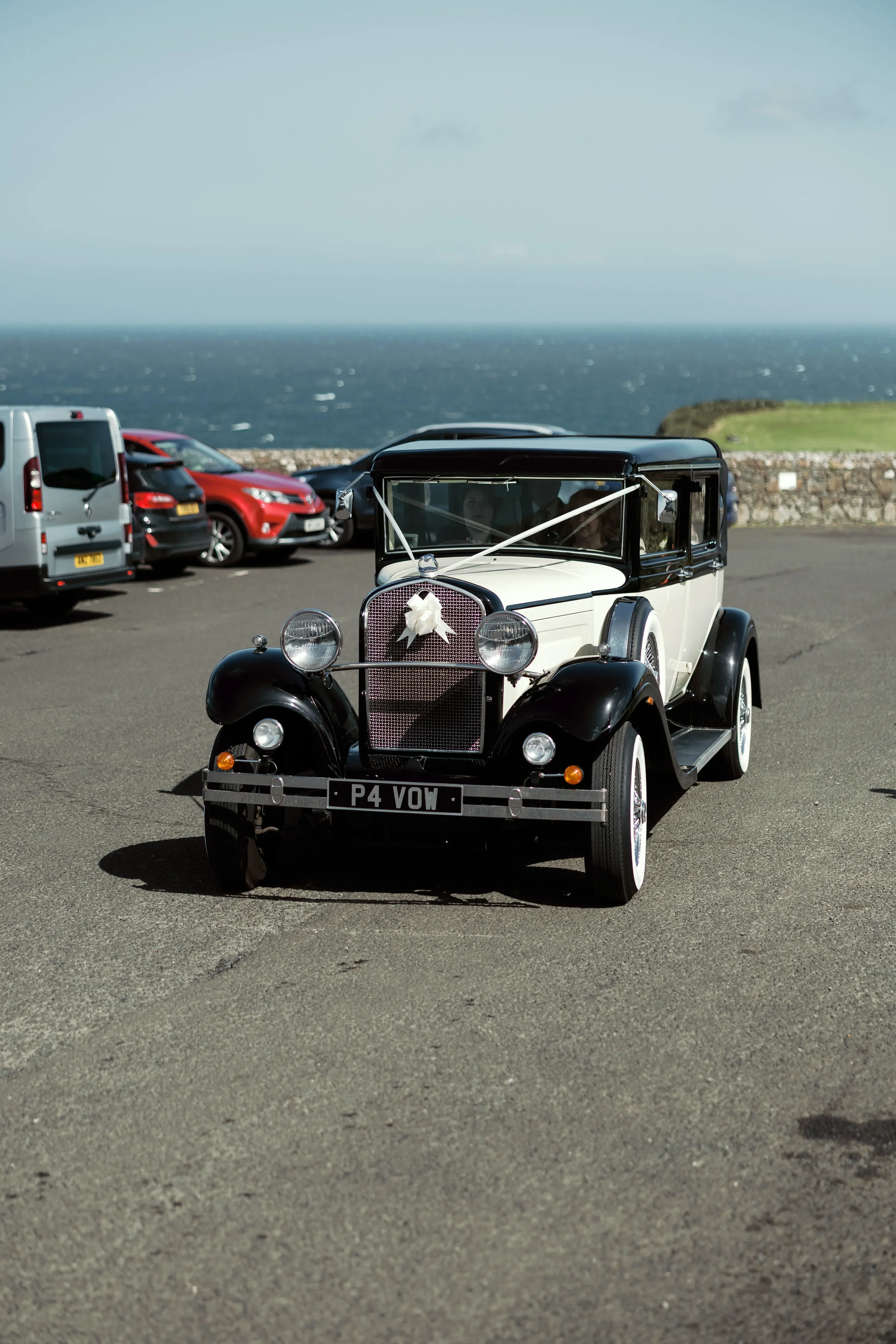 A vintage black and white car decorated with a white ribbon, likely for a wedding, parked near a seaside with the ocean in the background and other modern cars nearby.