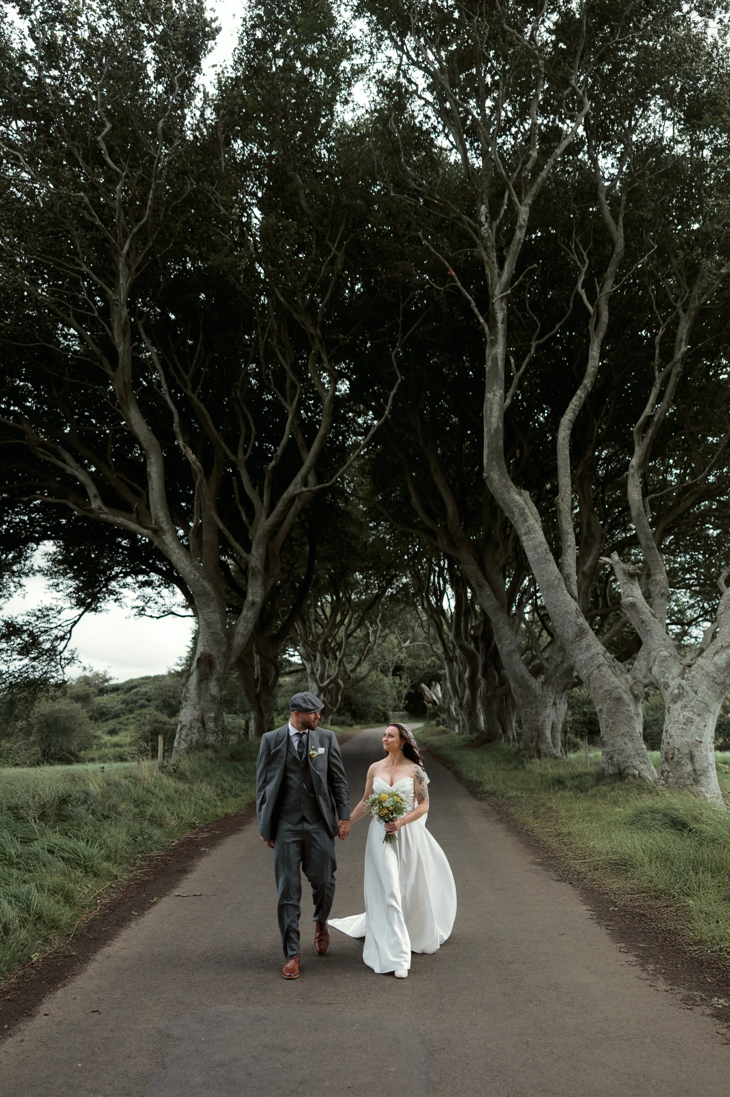 A bride and groom walking hand in hand down a tree-lined road.