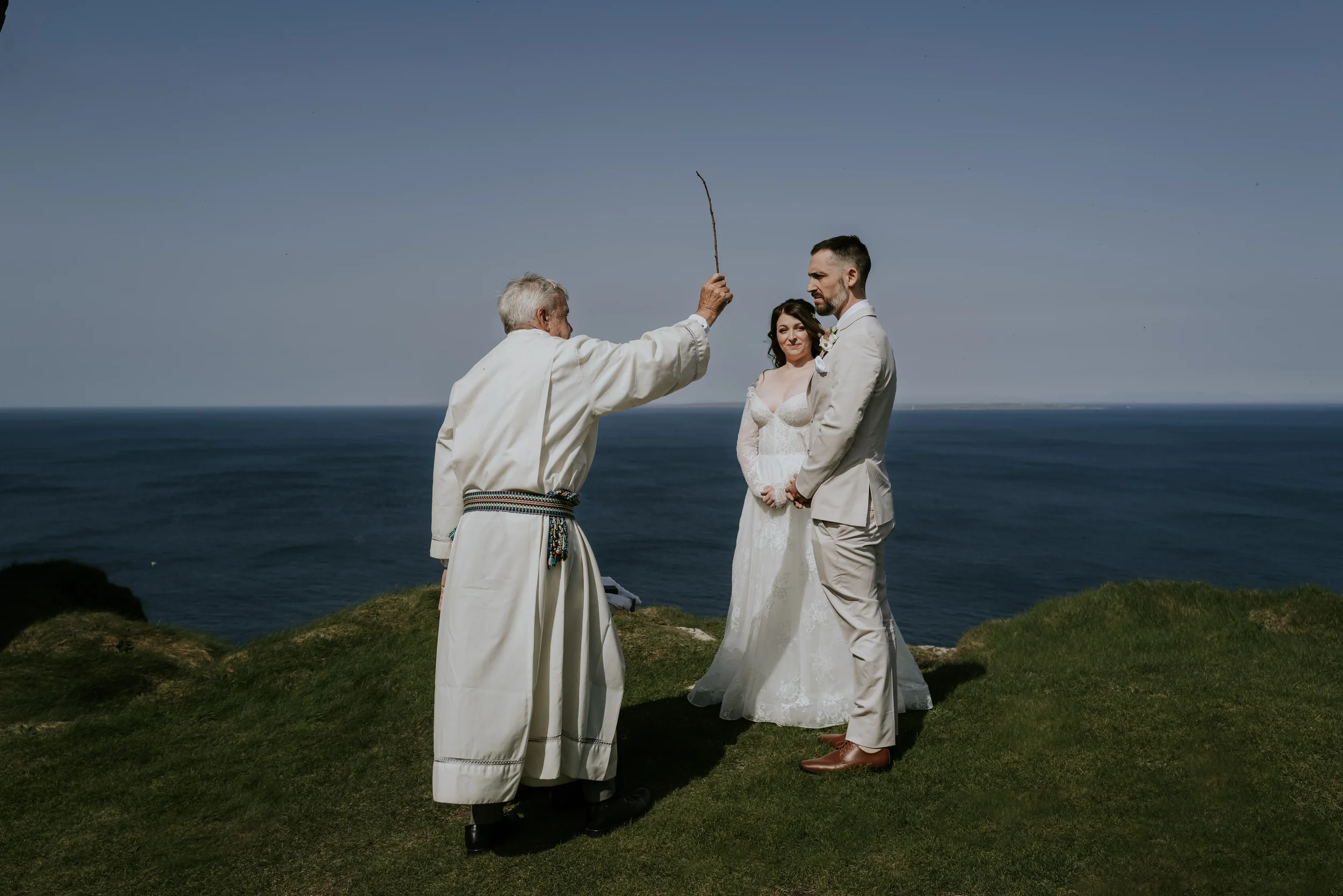 A bride and groom standing on grass near the ocean during a wedding ceremony, with a man in traditional attire holding a stick, possibly officiating the ceremony.
