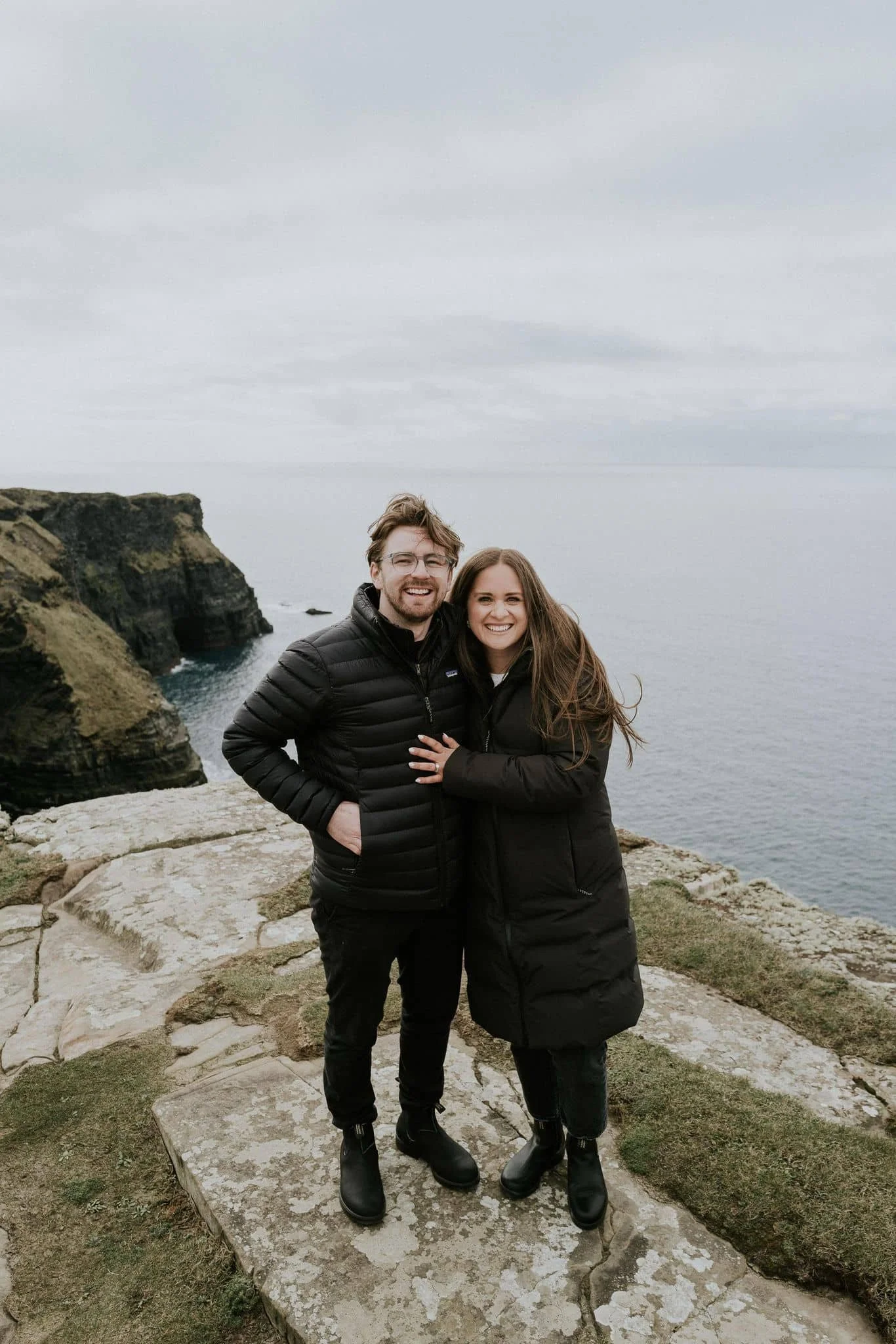 A smiling couple in black jackets standing on a stone ledge near the coast, with cliffs and the ocean in the background