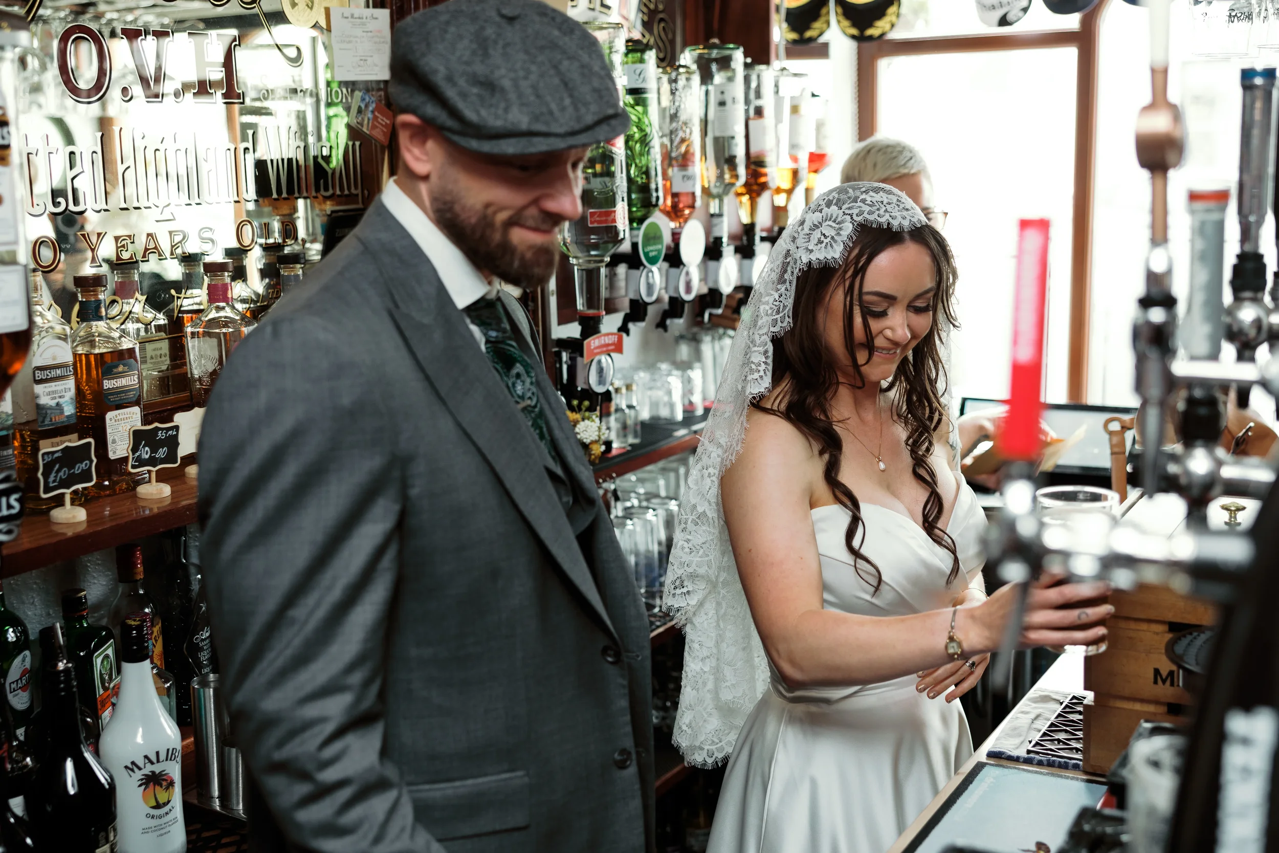 A man and woman dressed in wedding attire standing at a bar, smiling.