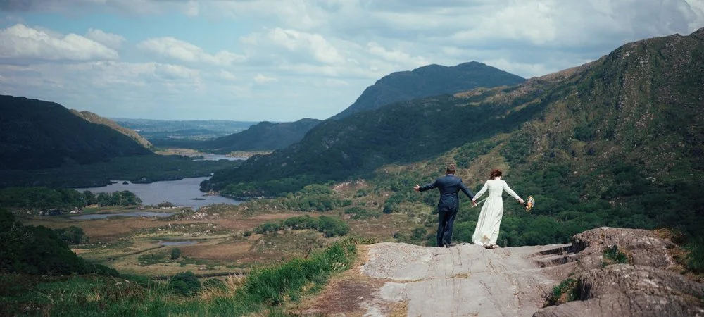 A newlywed couple holding hands and walking on a rocky trail in a scenic mountainous landscape with a river in the distance after their Ireland wedding