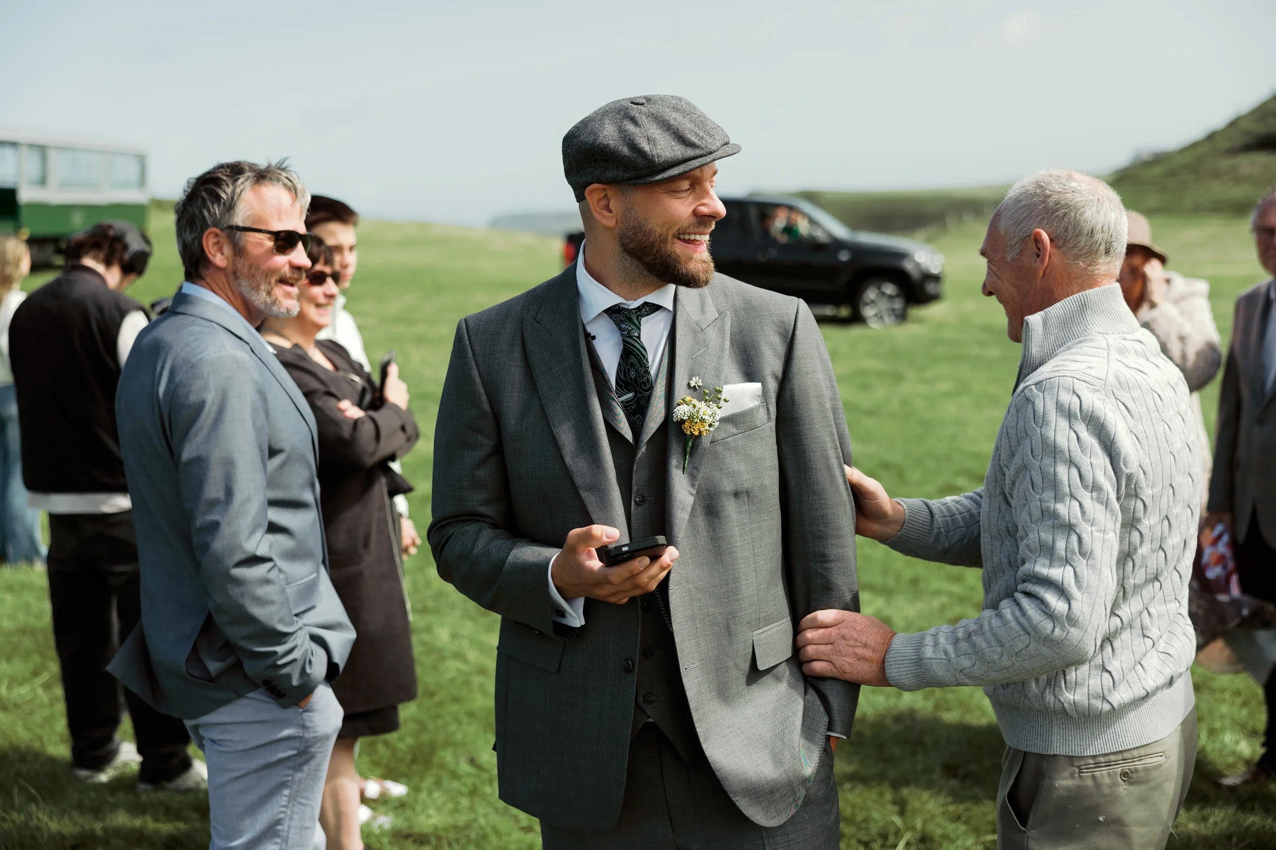 A man in a gray suit with a gray newsboy cap, holding a phone, smiling as an older man in a light gray sweater touches his arm at an outdoor gathering on a grassy field with several people, cars, and rolling hills in the background.