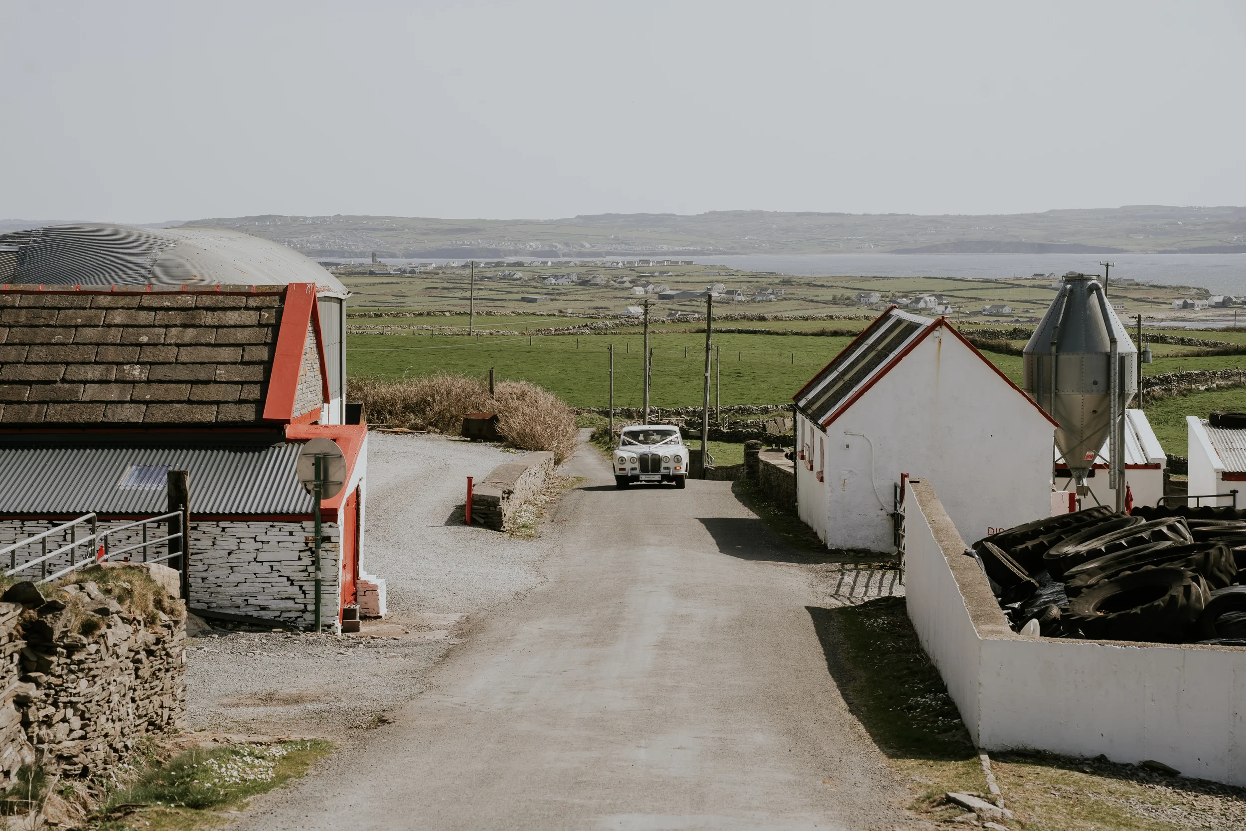 Dirt road in a rural area with white buildings, a vintage car, and green fields in the background under a cloudy sky.