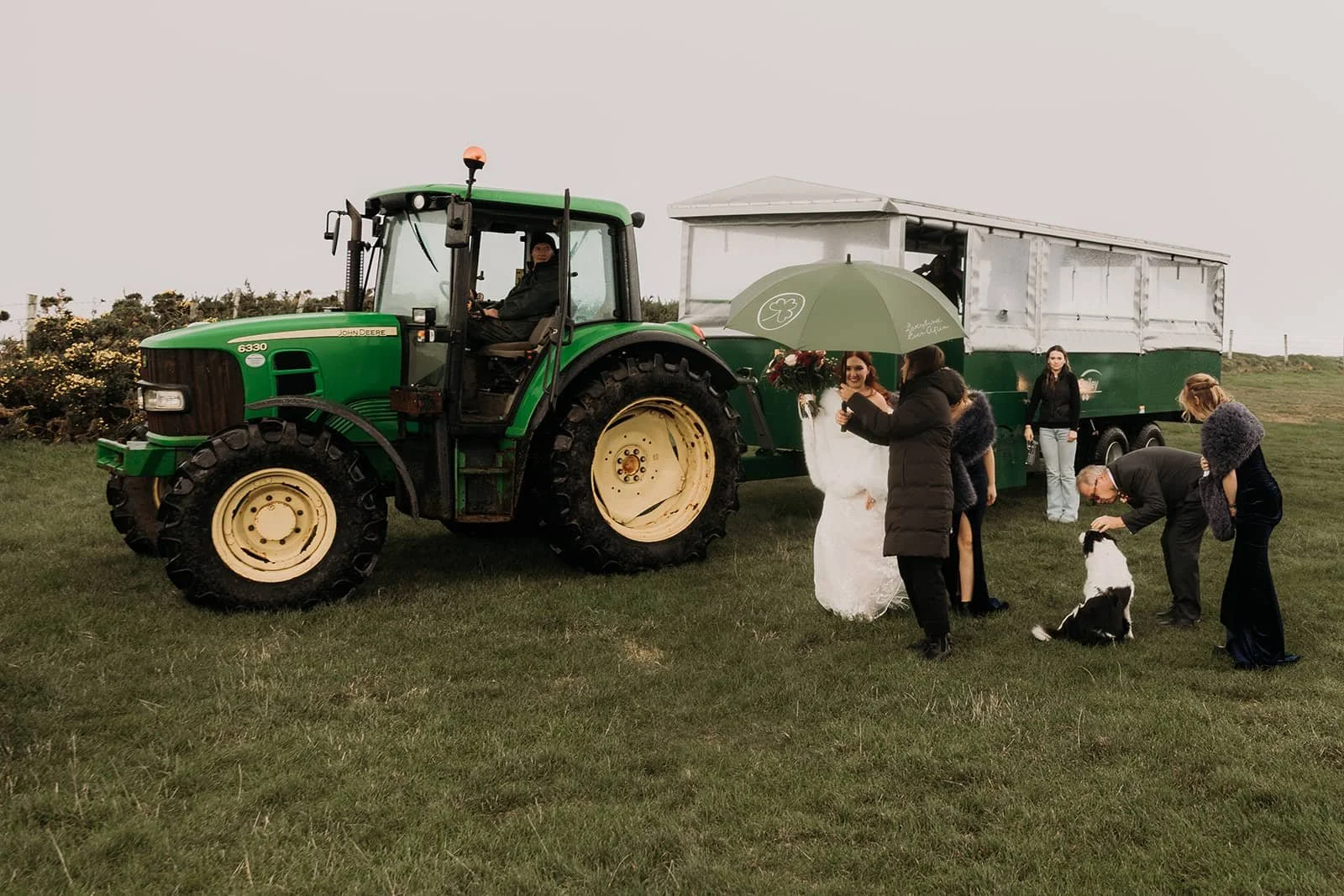 A group of people in wedding attire gather around a large green tractor on a grassy field at an Ireland wedding. Some are holding a green umbrella, while others are petting a black and white dog. In the background, there is a trailer connected to the