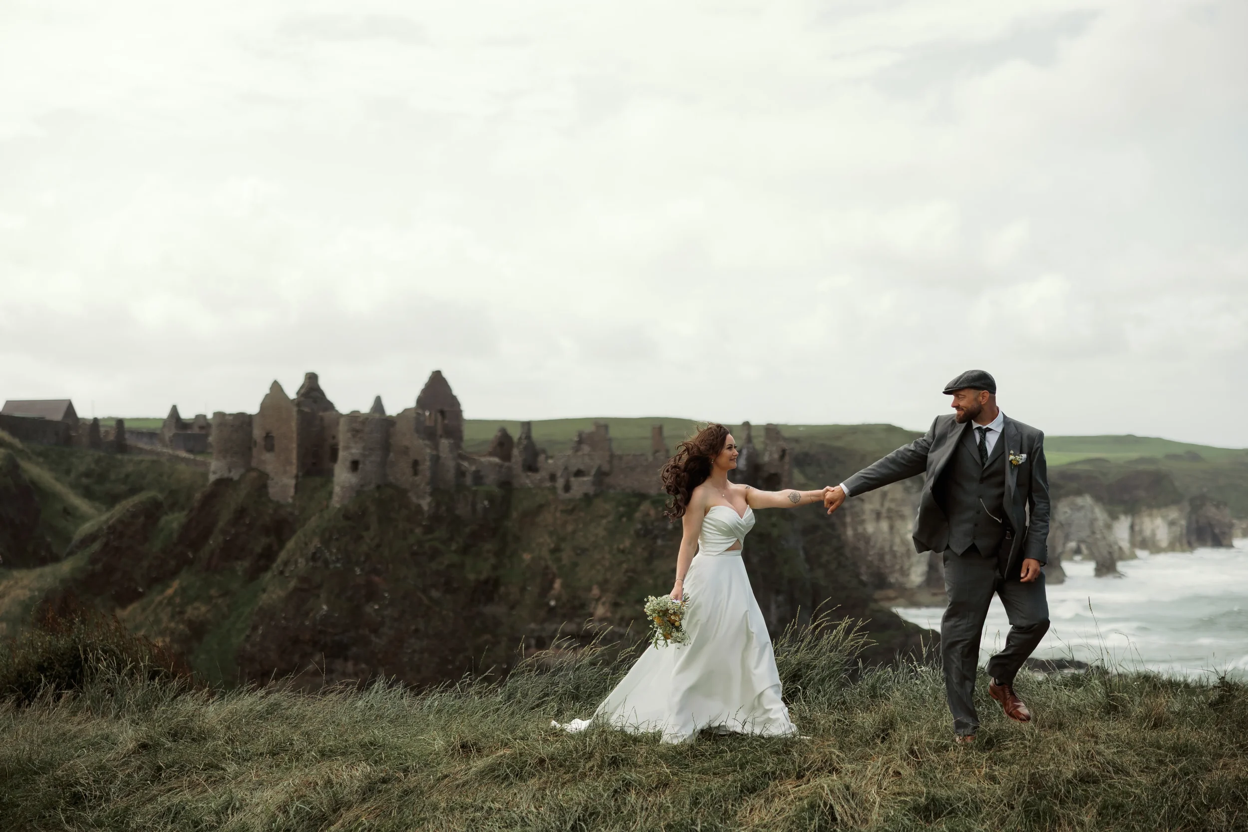 A bride and groom holding hands and dancing outdoors on a grassy area, with ruined castles on a hill and the ocean in the background.
