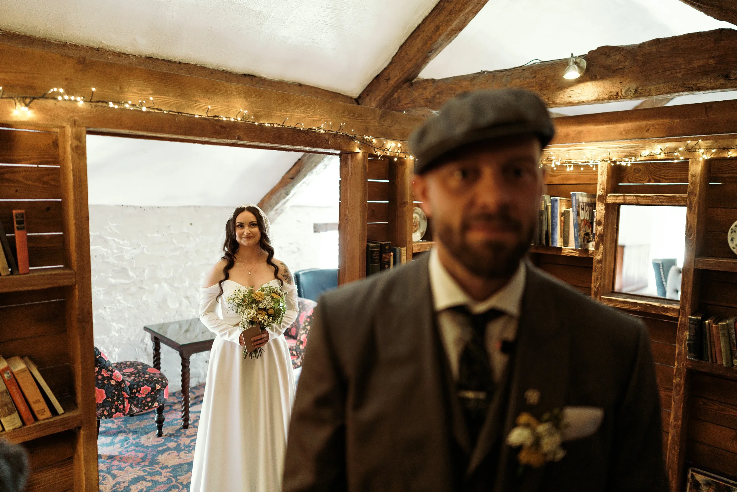 A bride with dark hair, wearing a white off-the-shoulder wedding dress, is standing inside a cozy, rustic room with white brick walls and wooden beams, holding a bouquet of flowers. A man in a suit and flat cap stands in the foreground, slightly out 