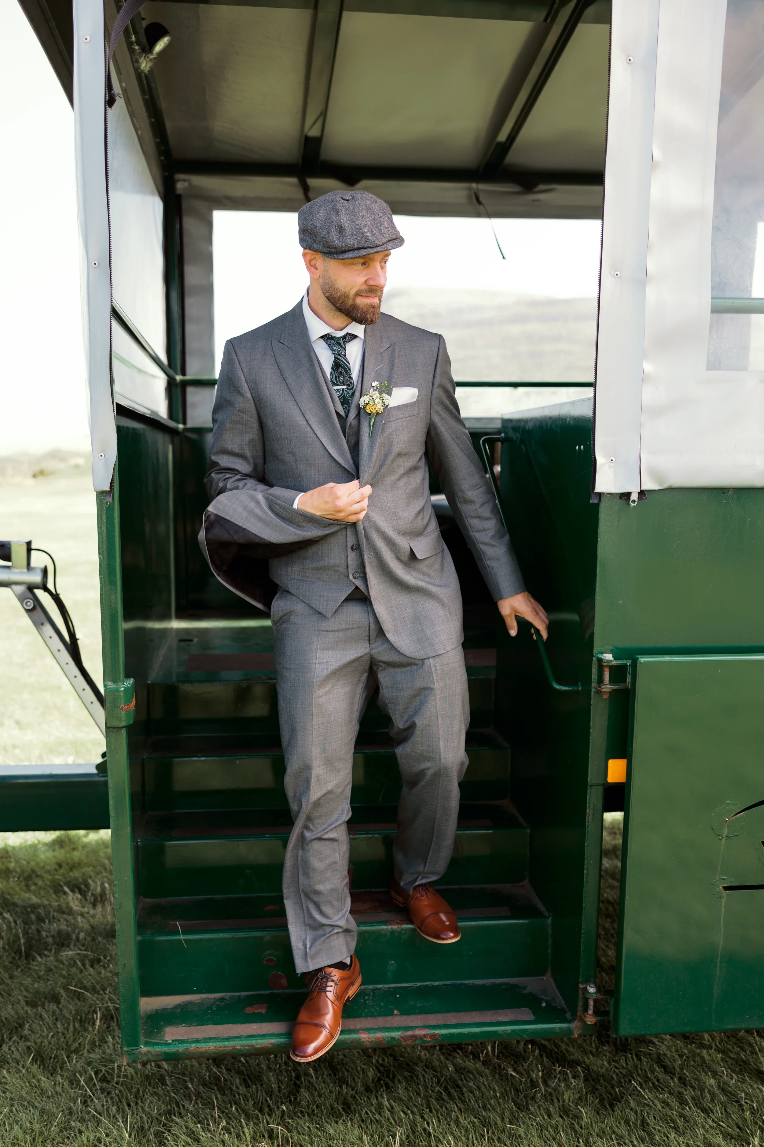 A man in a gray suit and a flat cap steps out of a green horse-drawn carriage onto a grassy field.