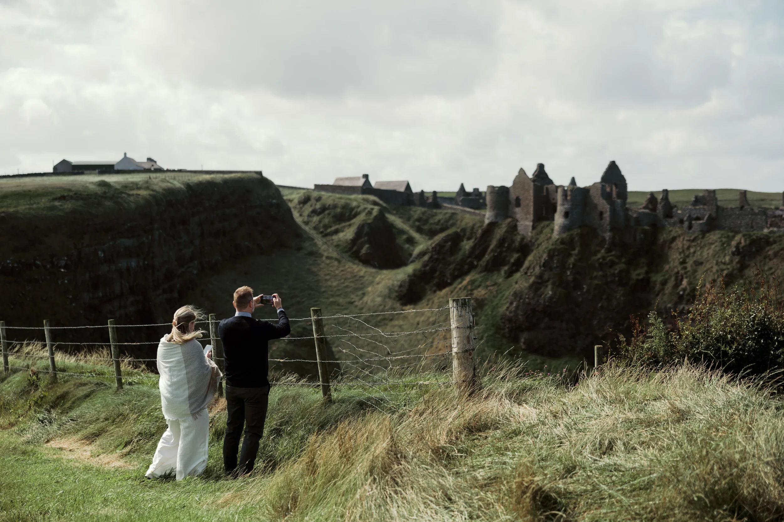 Two people, a man and a woman, standing near a fence on a grassy hill, taking photos of a historic castle ruins on a distant cliff under a cloudy sky.