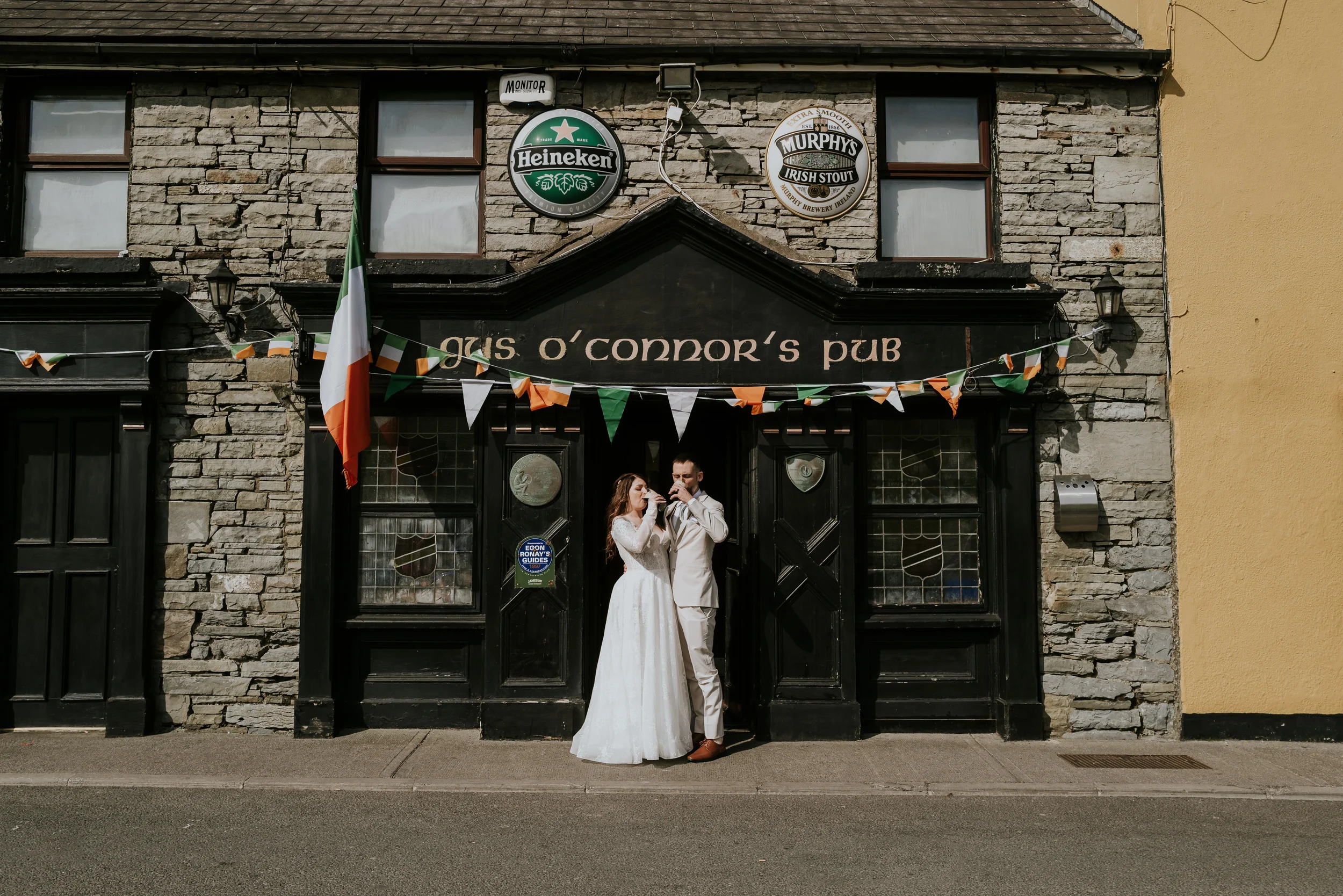 A bride and groom in wedding attire standing outside a pub named 'gris o'connor's pub' decorated with Irish flags and bunting, with signs for Heineken and Murphy's Irish Stout on the building.