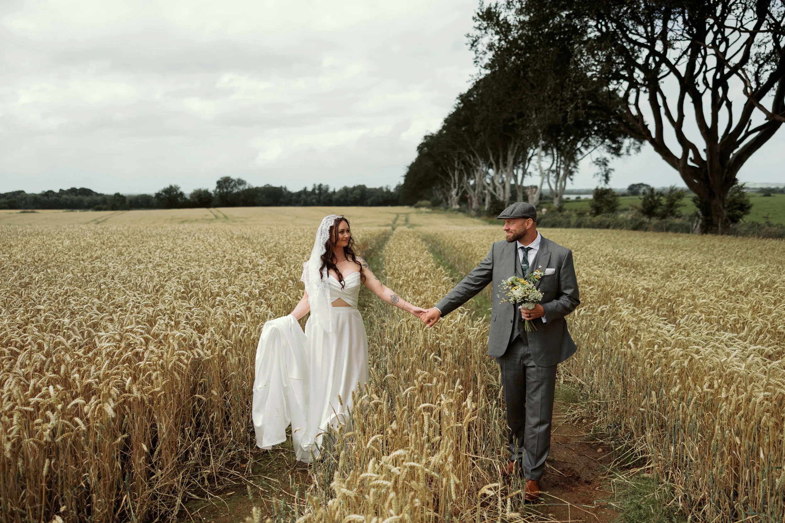 A bride and groom holding hands in a wheat field, with the bride in a white dress and the groom in a gray suit holding a bouquet, under a cloudy sky with large trees in the background.