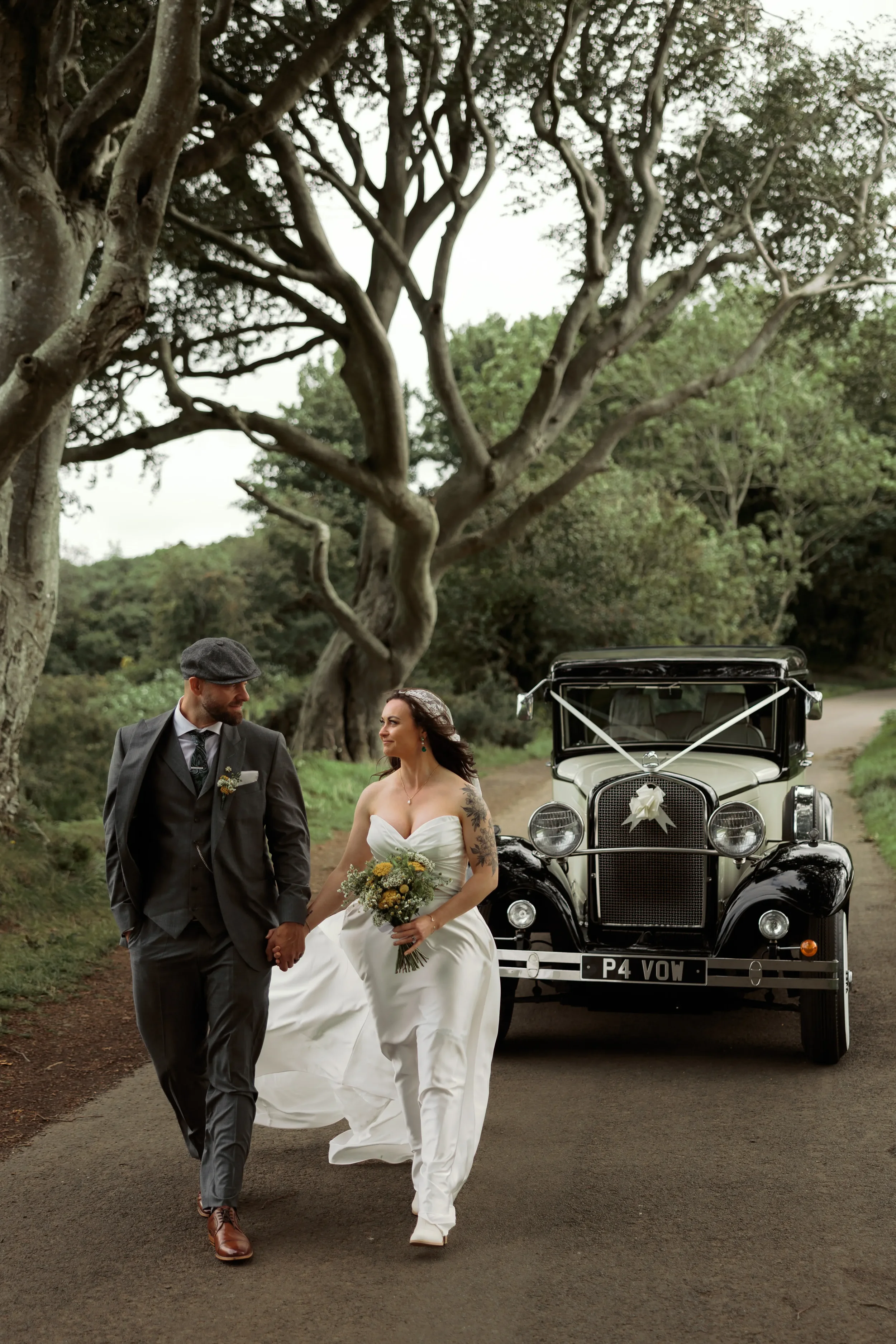 A newlywed couple holding hands walking in front of a vintage car on a dirt road surrounded by greenery and large trees, with the bride holding a bouquet.