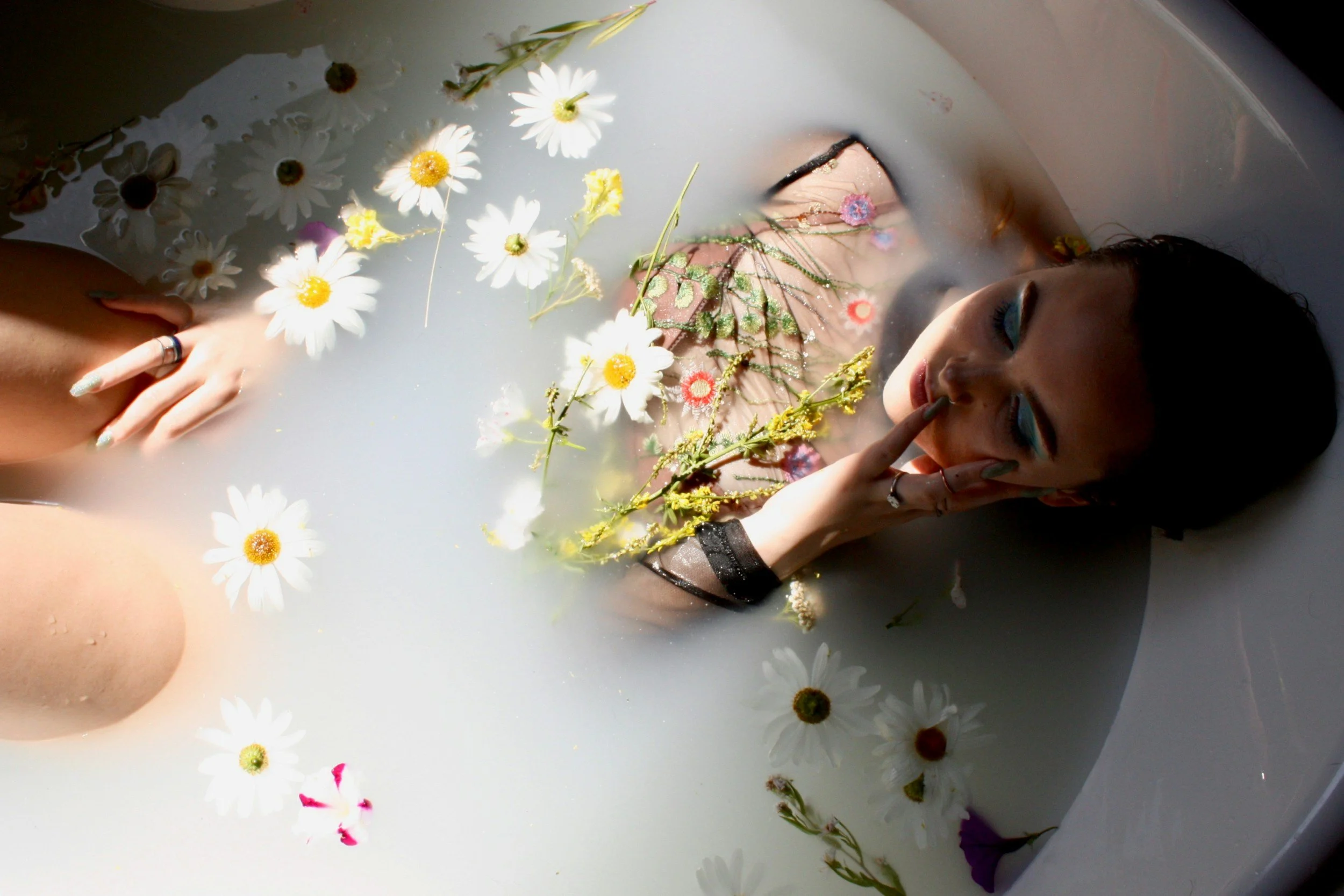A woman with blue eyeshadow and earrings is lying in a bathtub filled with milky water and floating daisies and flowers, touching her face with her hand.