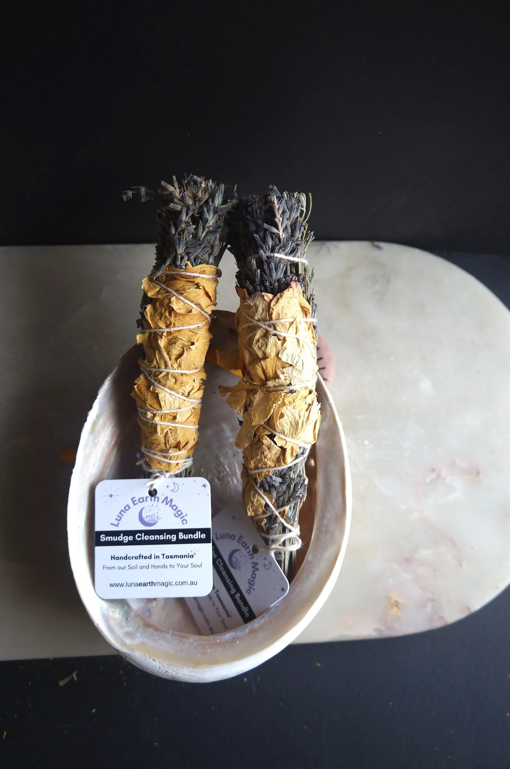 Dried lavender bundle tied with string resting on a white ceramic dish, with a label attached, beside yellow roses and green leaves on a black surface.
