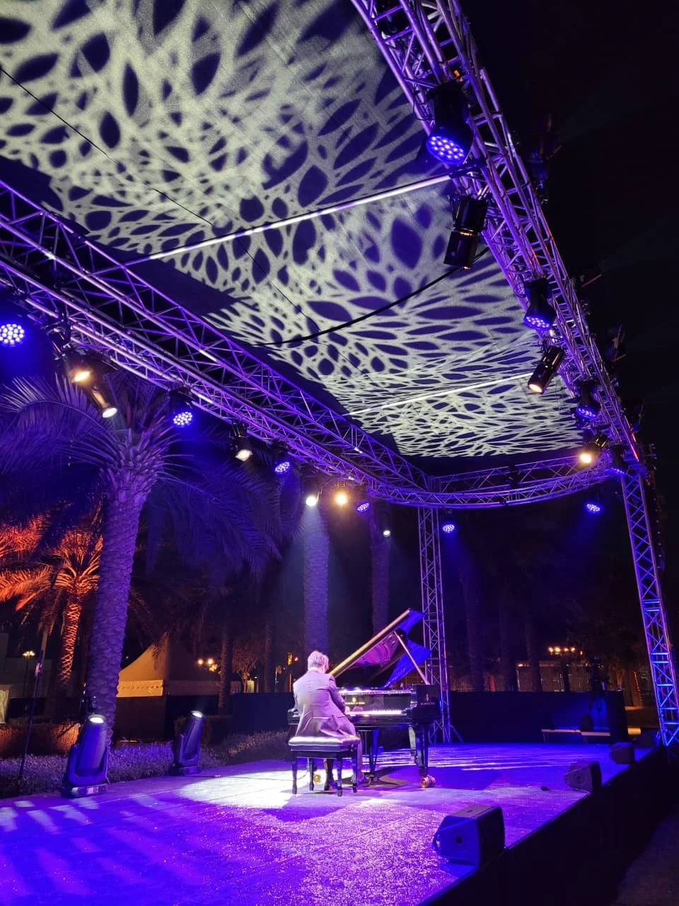 A person playing a grand piano on an outdoor stage under purple and yellow lighting, with palm trees in the background and a decorative pattern projected on the canopy overhead.
