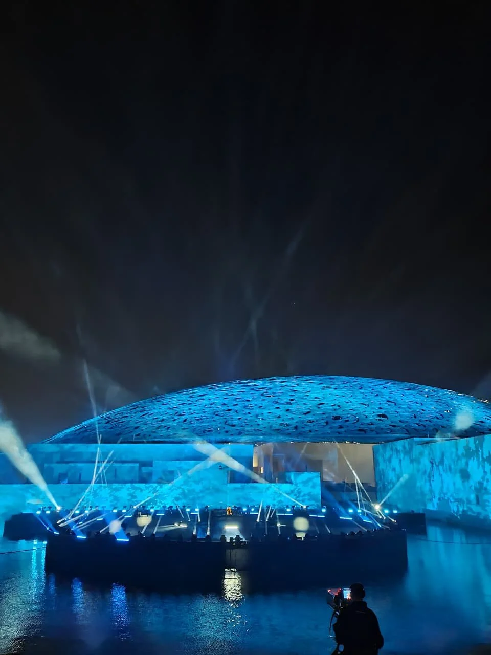 Nighttime scene of a modern building with a dome-shaped roof illuminated in blue, with bright spotlights and projections on the structure, reflecting in a body of water in the foreground. A person in dark clothing stands in the water taking a photo of the scene.