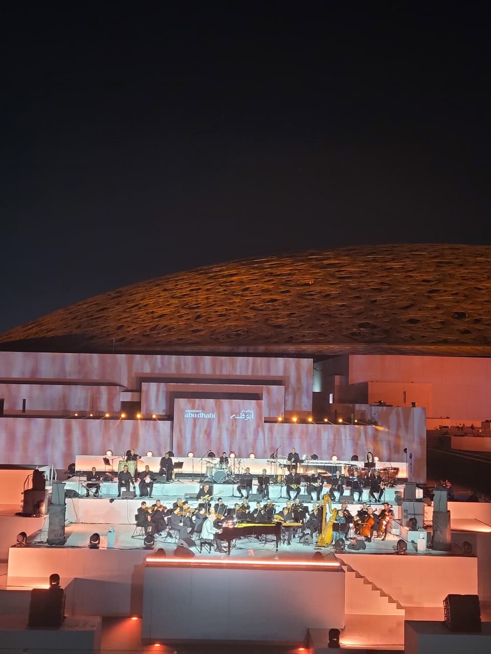 A nighttime outdoor concert featuring a large orchestra on a multi-tiered stage with musicians, a grand piano, and a harp, set against a modern building with an illuminated, curved rooftop.