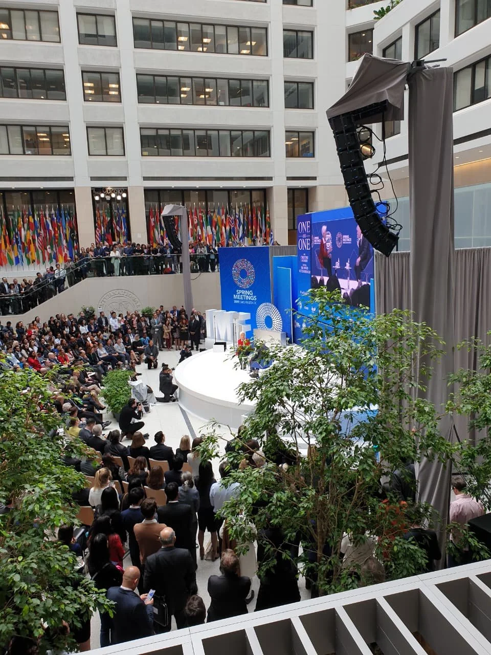 A large indoor conference with a stage, screens, and numerous attendees seated and standing, at the United Nations headquarters during a spring meeting.