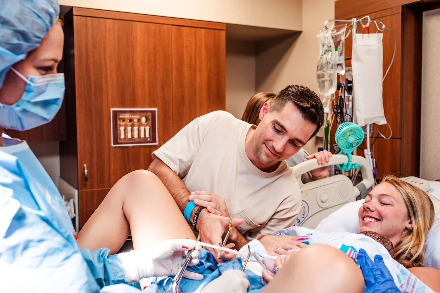 Hospital birth proud dad cuts the cord with mom smiling on.