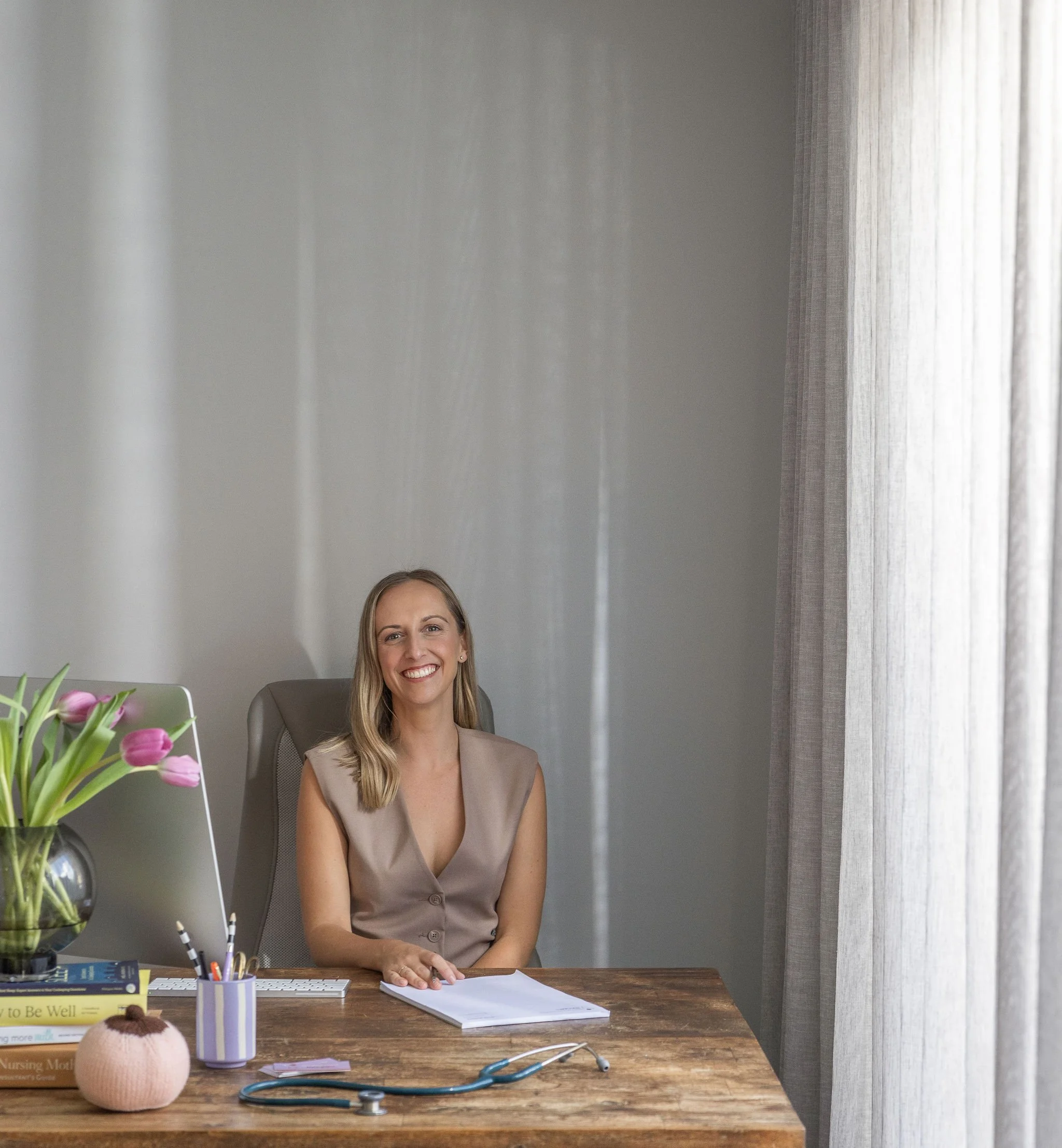 A smiling woman sitting at a wooden desk with medical and office items, including a stethoscope, notebooks, a pen holder with pens, pink tulips in a vase, and books, in a room with gray walls and curtains.