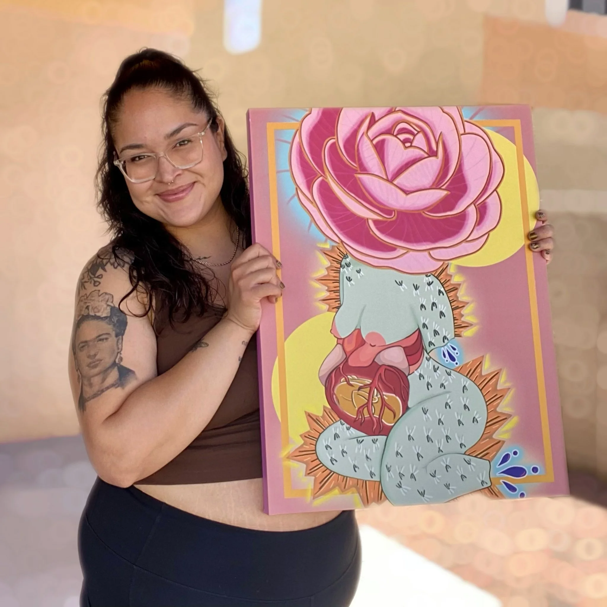 Woman with glasses and tattoos holding a colorful painting of a cactus with a large pink rose on top, smiling.