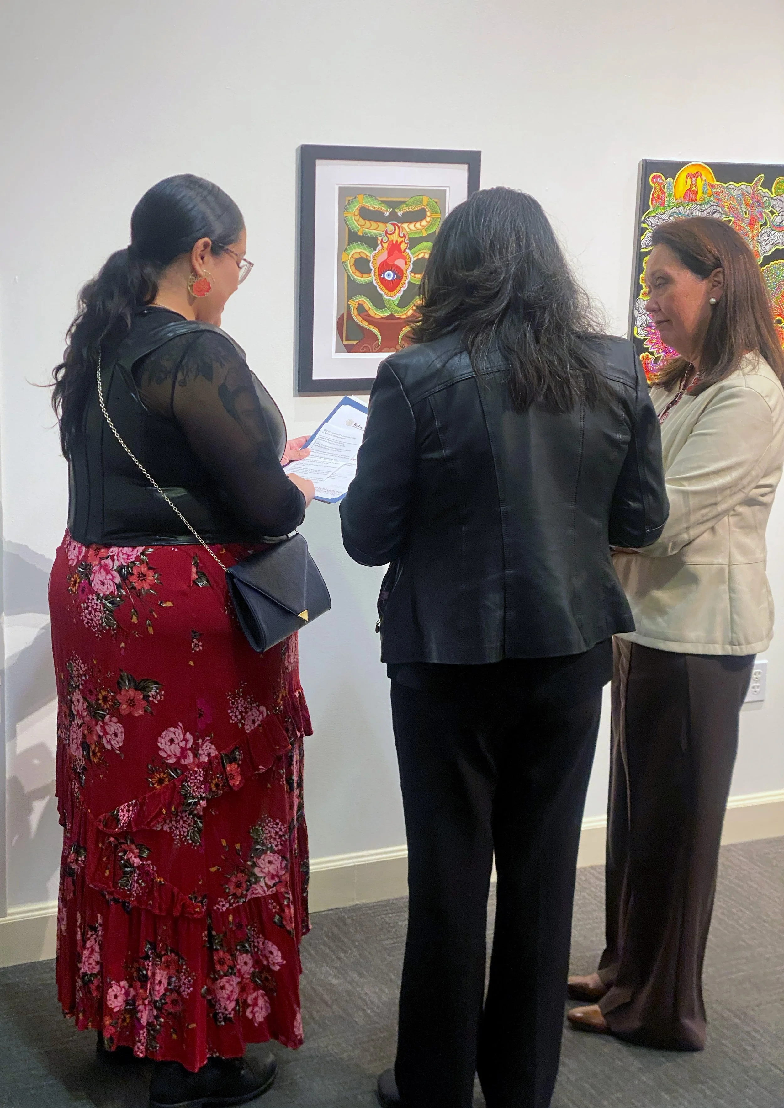 Three women standing in front of artwork at an art gallery, engaged in conversation, with one woman holding a brochure.