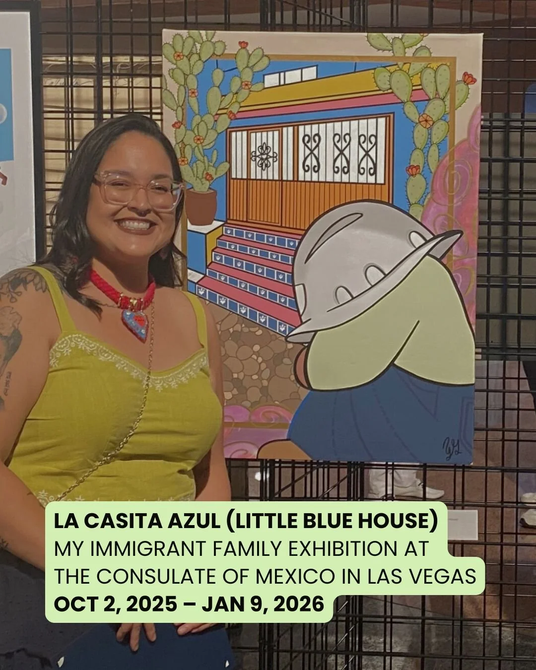 A woman wearing glasses, a yellow top, and red jewelry stands next to a colorful artwork depicting a house, potted cacti, and a cartoon character with a hat covering its face, all behind a black wire grid. The woman is smiling and has tattoos on her 