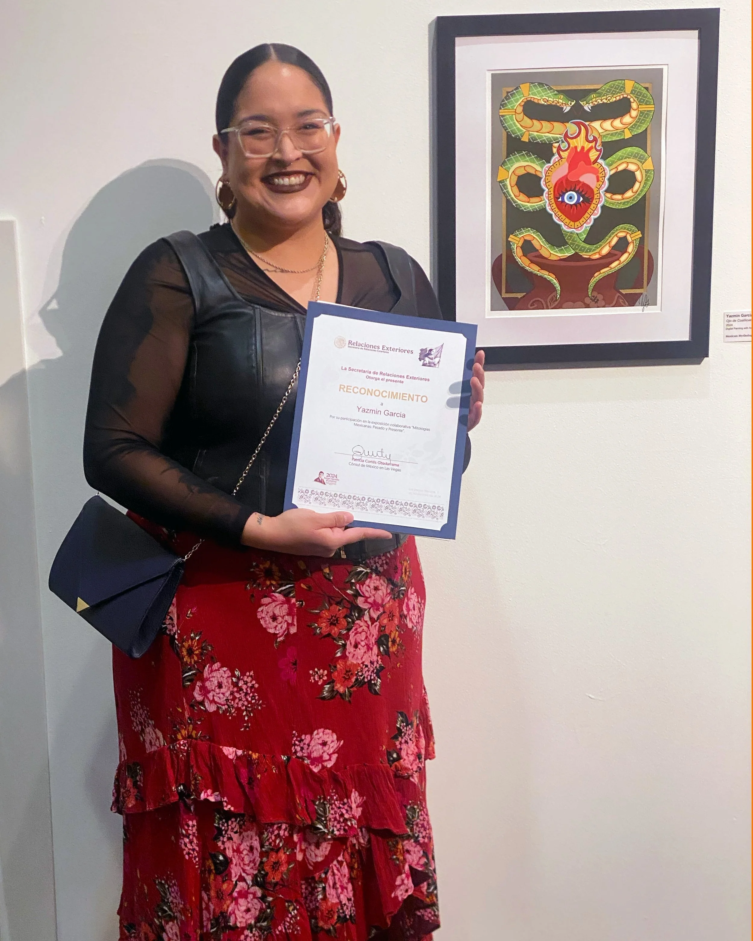 A woman with glasses and dark lipstick holding a certificate while standing in front of a framed artwork of a snake and a dragon with an eye in the middle of a fire.