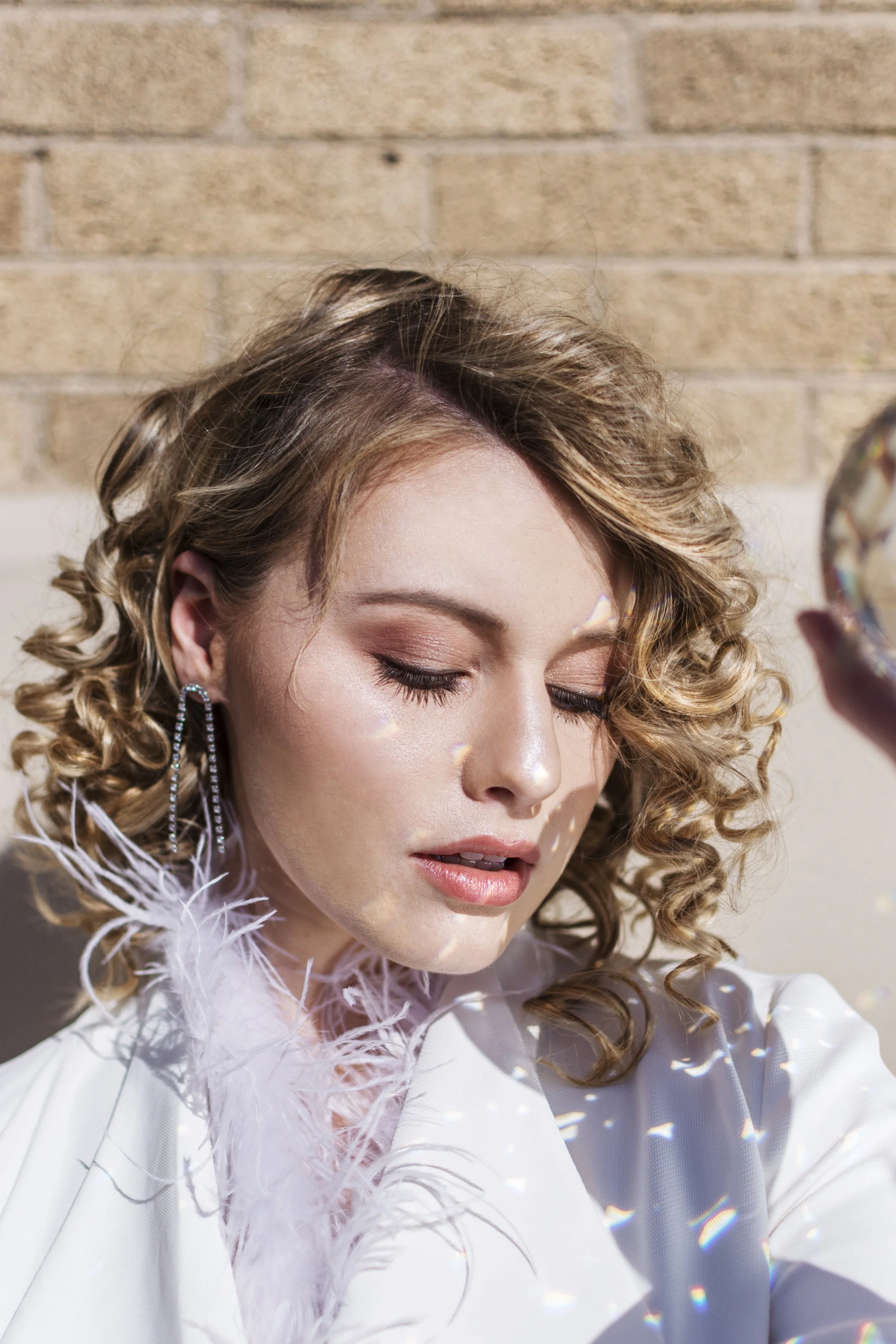 A woman with curly blonde hair and earrings, wearing a white outfit with feather details, looking down with closed eyes against a brick wall. Shot by Mary Huong.