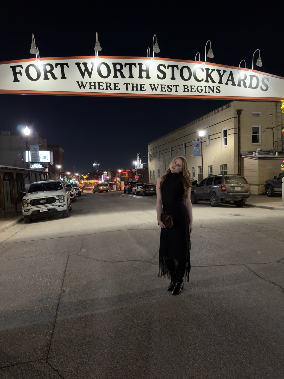 A woman standing on a city street at night, with a large sign overhead reading "Fort Worth Stockyards" and "Where the West Begins" in the background. The street is lined with parked cars and buildings, and the scene is illuminated by streetlights and signs.