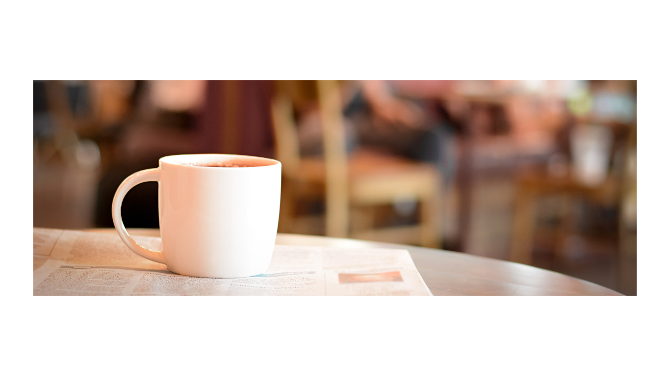 A white ceramic mug filled with hot coffee or tea on a table covered with a newspaper, with a blurred cafe background.