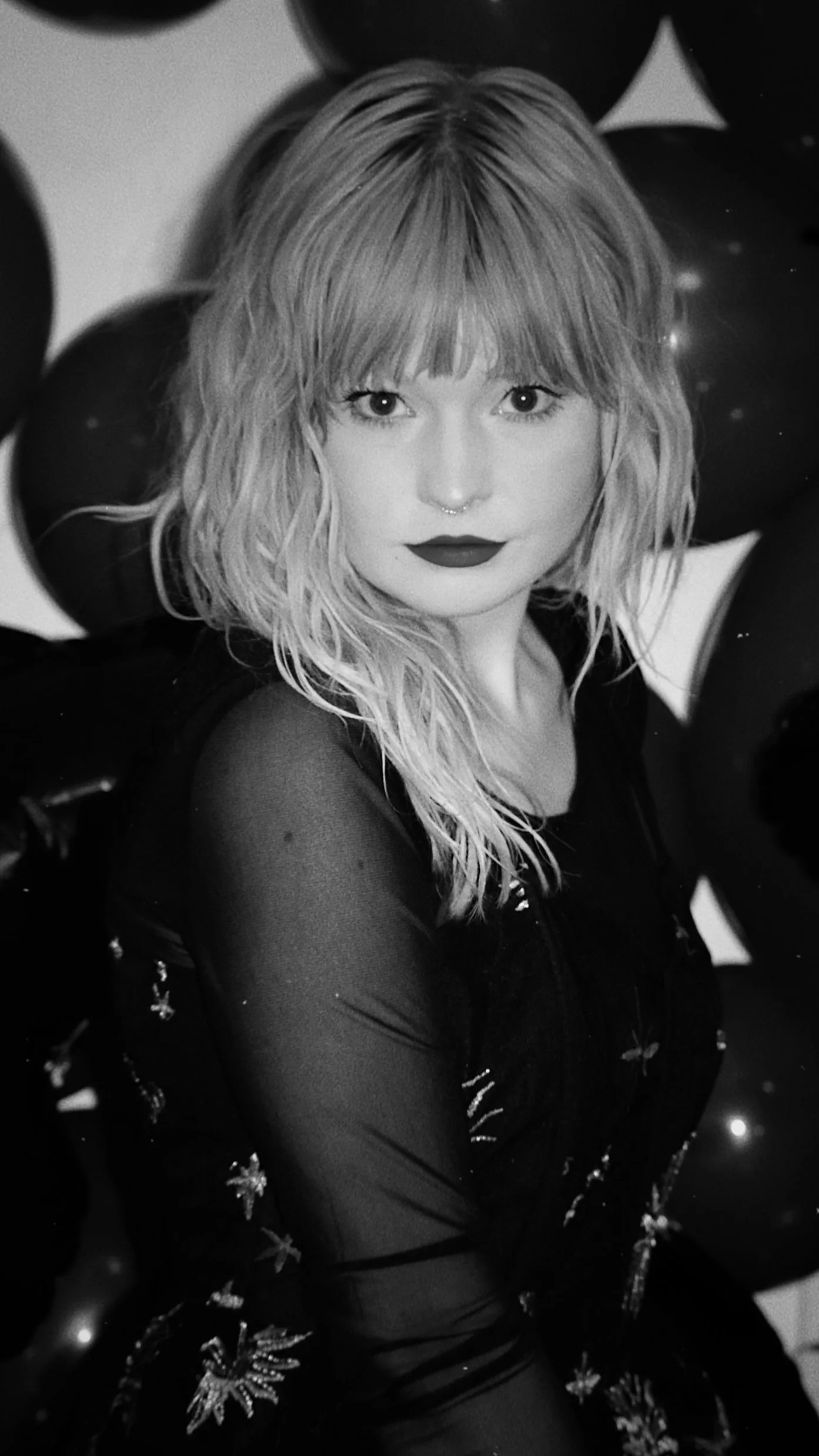 A black and white portrait of a woman with wavy blonde hair and bangs, wearing dark lipstick and a dark sheer top with embroidered patterns, surrounded by black balloons.
