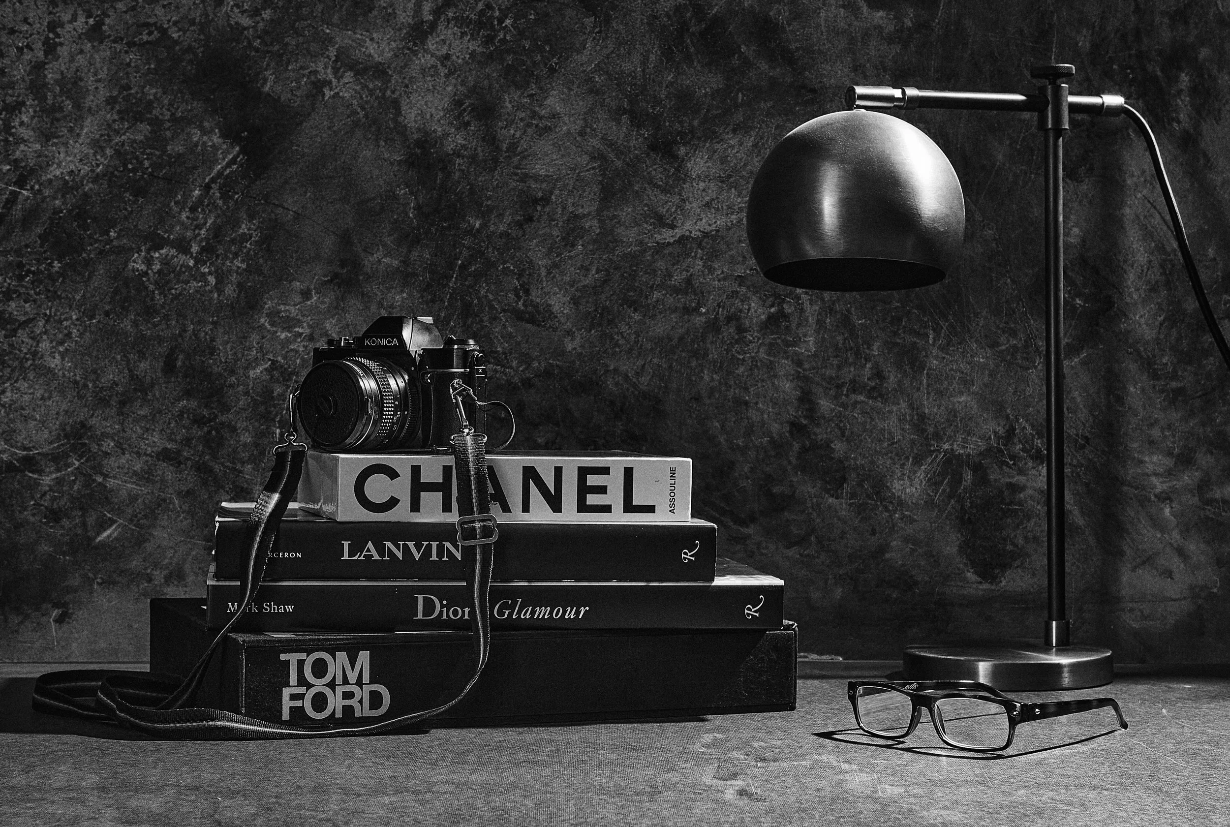 Black and white photo of a stack of fashion and luxury brand books, with a vintage camera on top, placed beside a modern desk lamp and a pair of reading glasses on a textured surface.