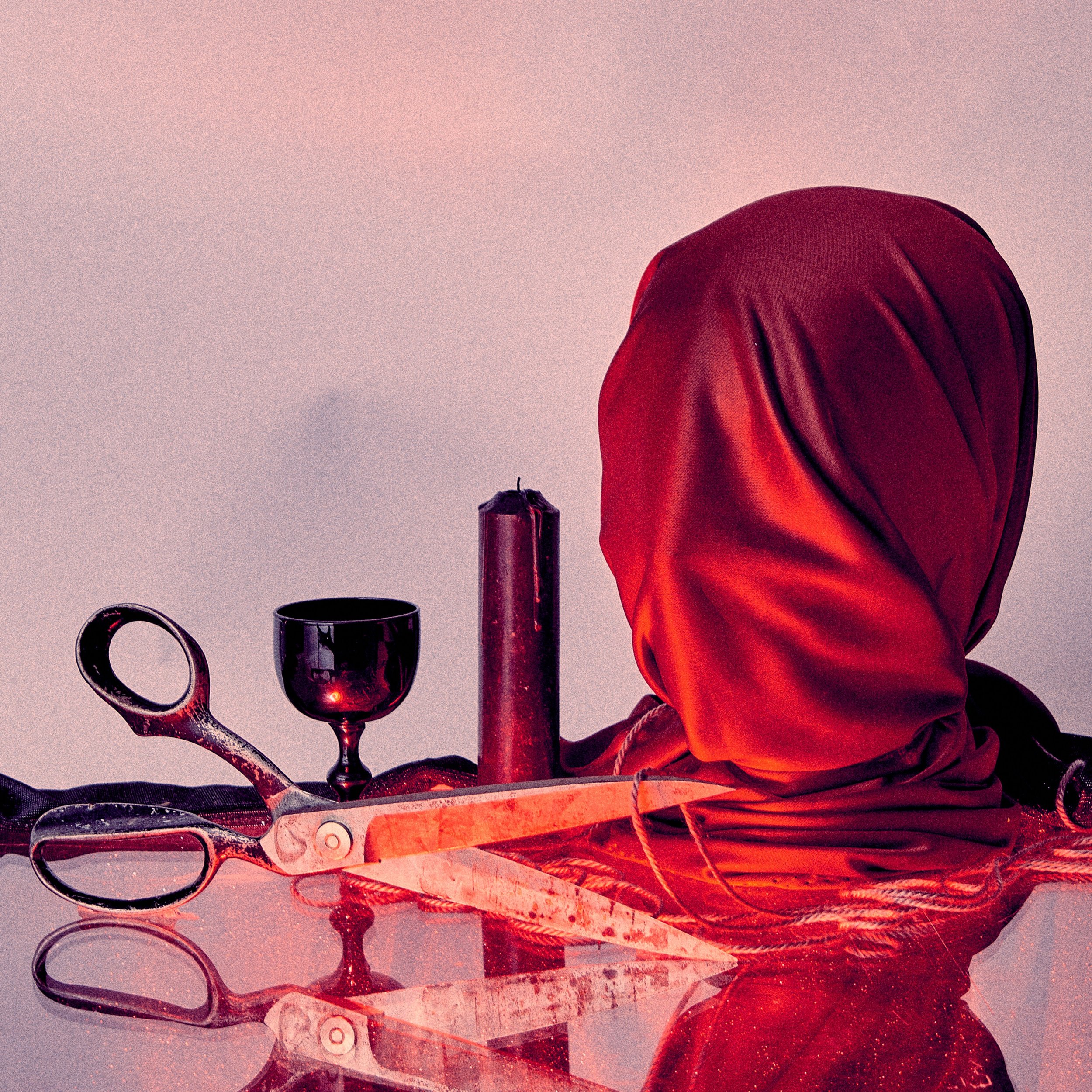A red cloth covering a human head, surrounded by a candle, a goblet, a pair of scissors, and a candle holder, all on a reflective surface with a plain background.