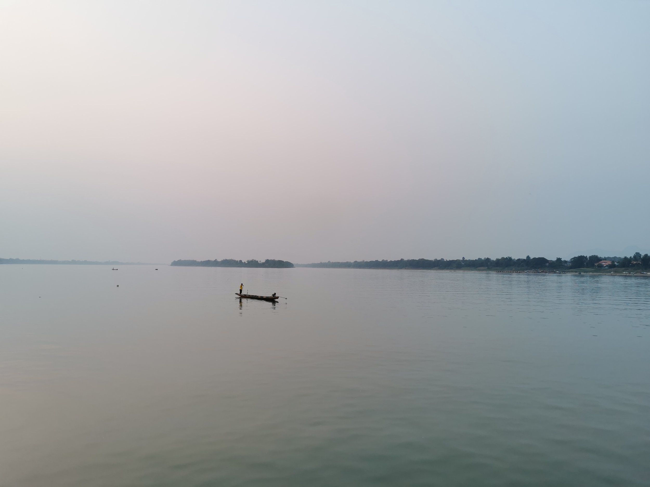 A calm body of water with a small boat containing a person, and an island or landmass in the background under a mostly cloudy or foggy sky.