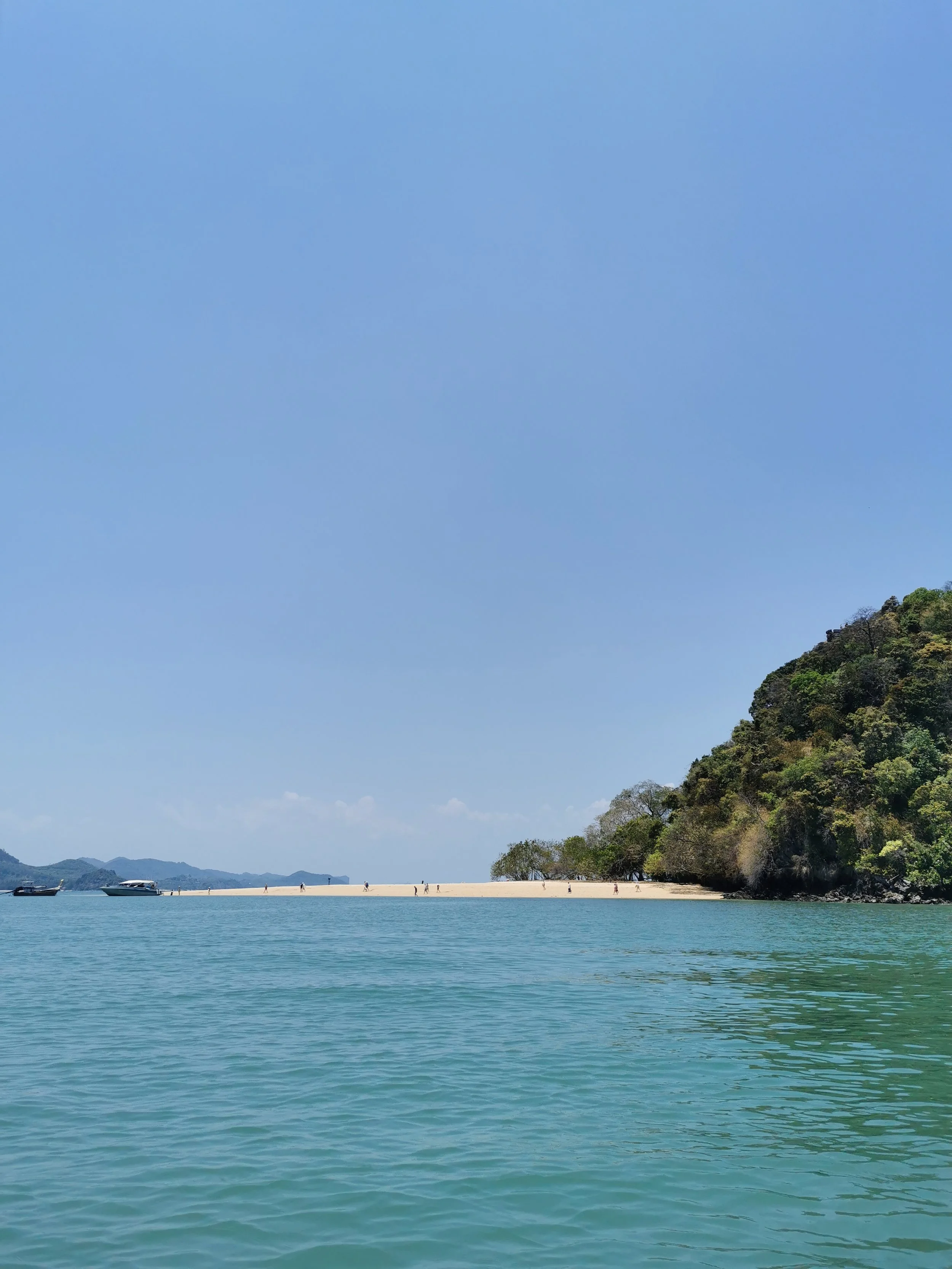 Calm blue water in the foreground, a sandy beach with a few people, lush green trees on a hillside, and a clear blue sky with some clouds.