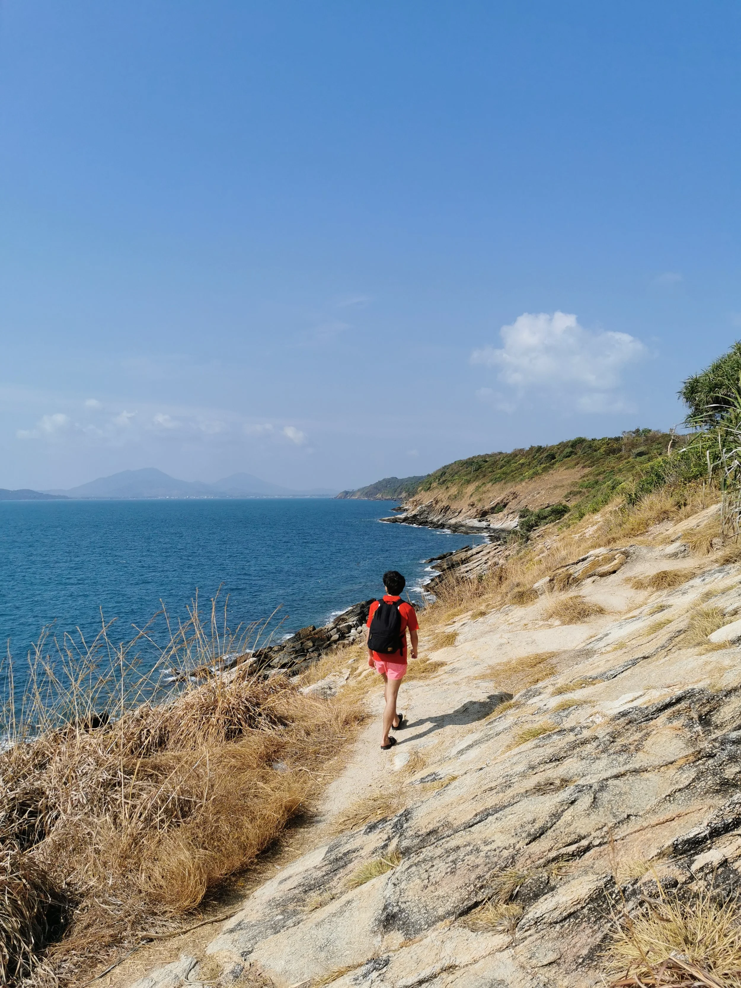 A person wearing red shorts and a red backpack walks along a rocky coastal path with dry grass on the sides. The path runs alongside the water, with hills and a distant mountain in the background under a blue sky with a few scattered clouds.
