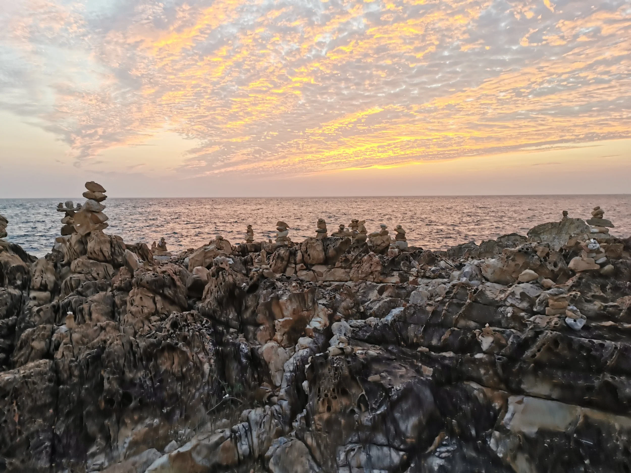 A rocky shoreline with stacked rocks in the foreground, a calm ocean extends to the horizon, and a sky filled with scattered clouds illuminated by a setting or rising sun.