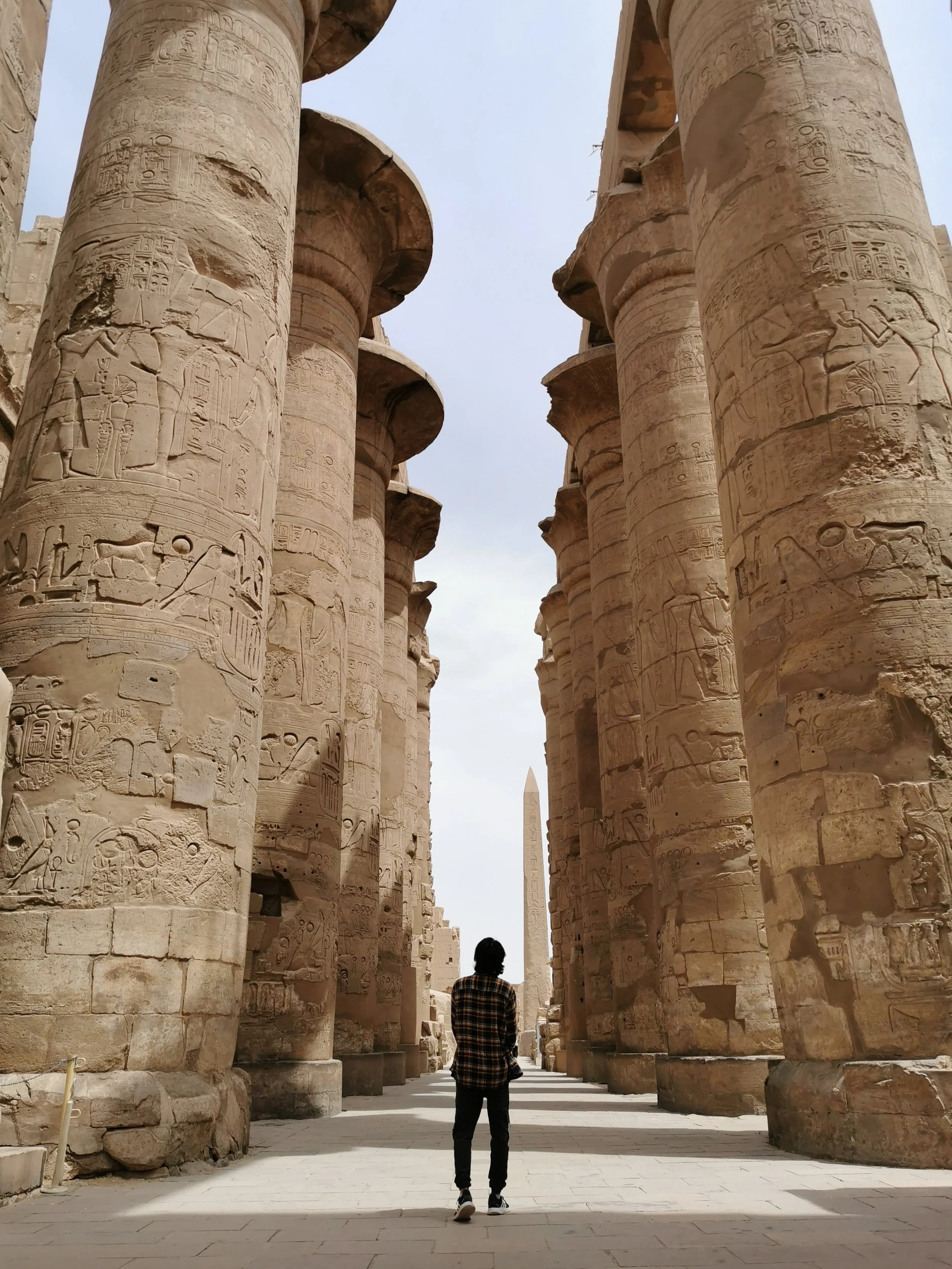 Person standing among large ancient Egyptian stone columns with detailed hieroglyphics, with the obelisk visible in the background.