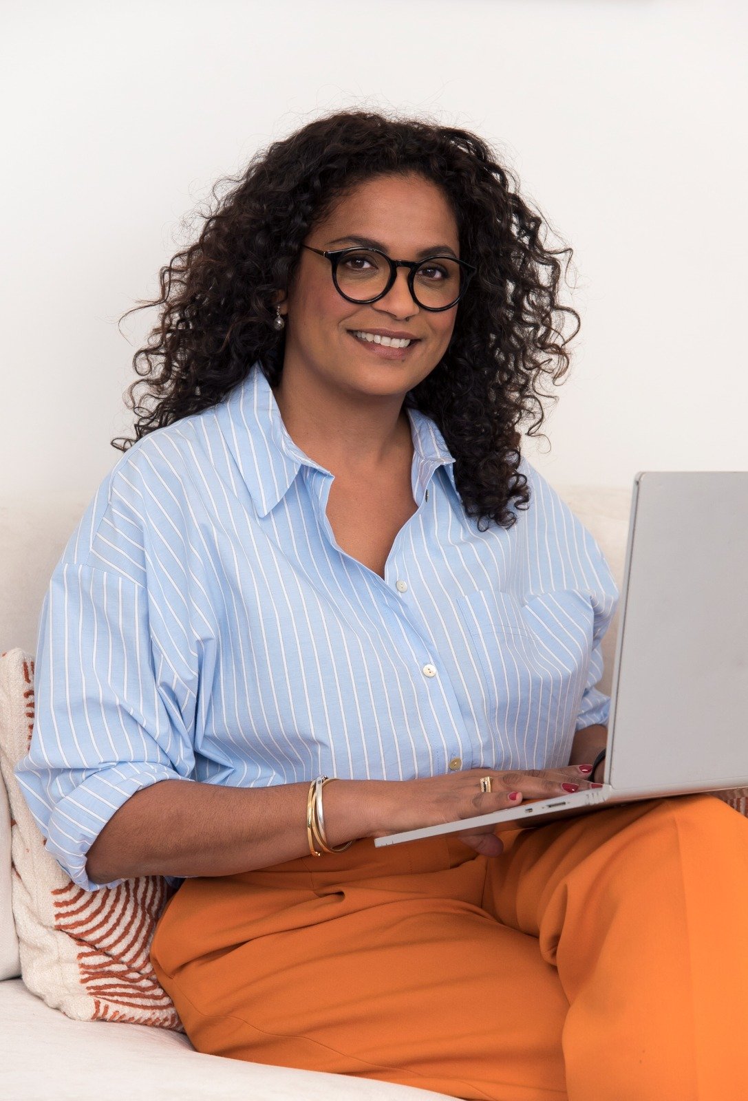 Une femme souriante avec des cheveux bouclés, portant des lunettes, une chemise à rayures bleues et blanches, et un pantalon orange, assise sur un canapé avec un ordinateur portable.