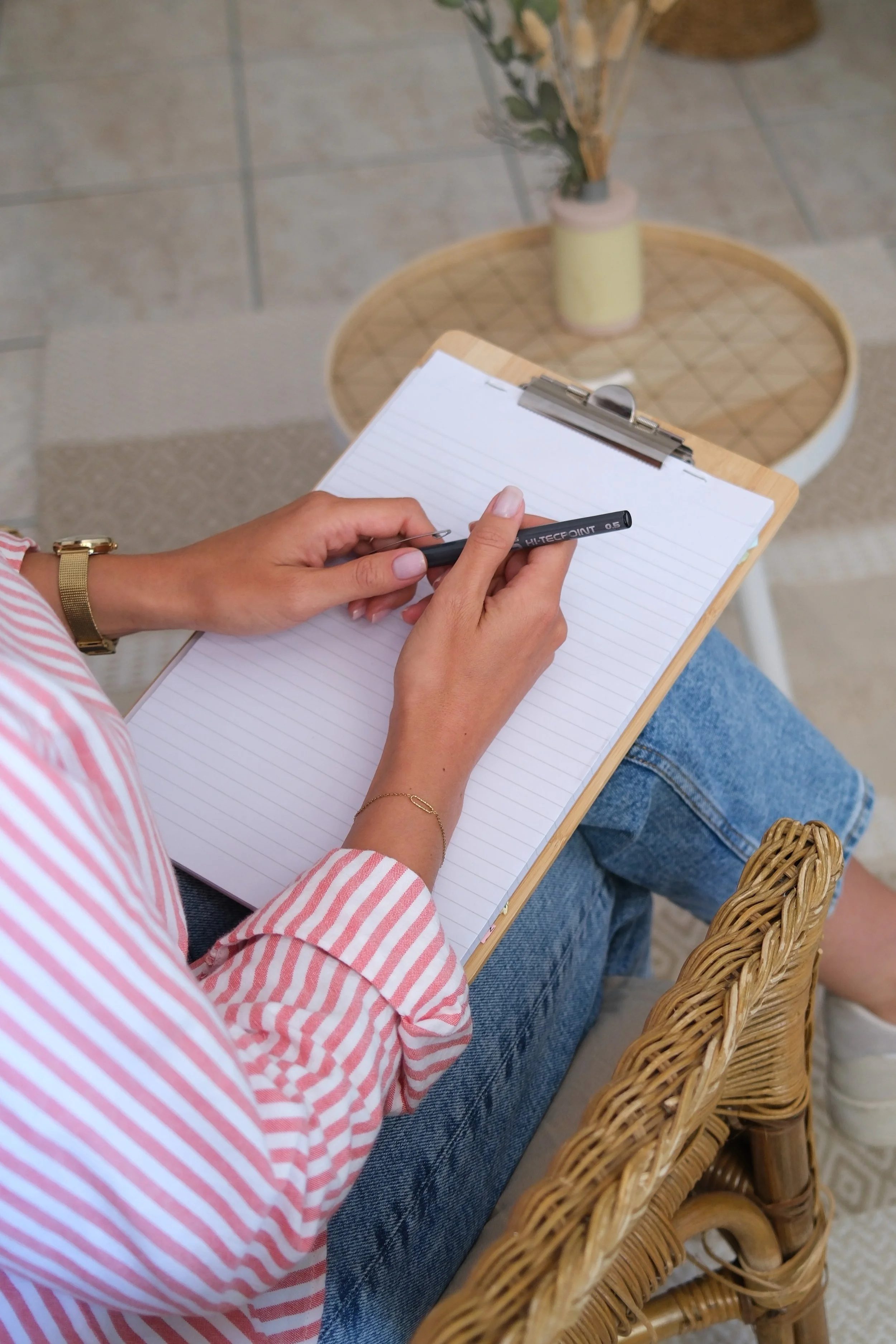 Une personne écrit sur une feuille de papier blanche avec un stylo noir, assise sur une chaise en rotin, portant une chemise à rayures rouges et blanches, un bracelet en or, et un jean. Il y a une petite table avec un vase de fleurs derrière elle.
