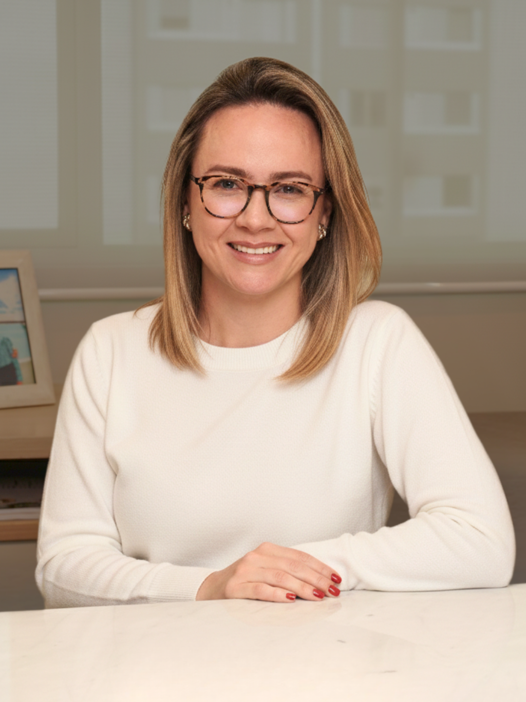 Nutrióloga con gafas, cabello castaño claro y sonrisa sentada en una mesa blanca con fondo de ventanas y una foto enmarcada a la izquierda.