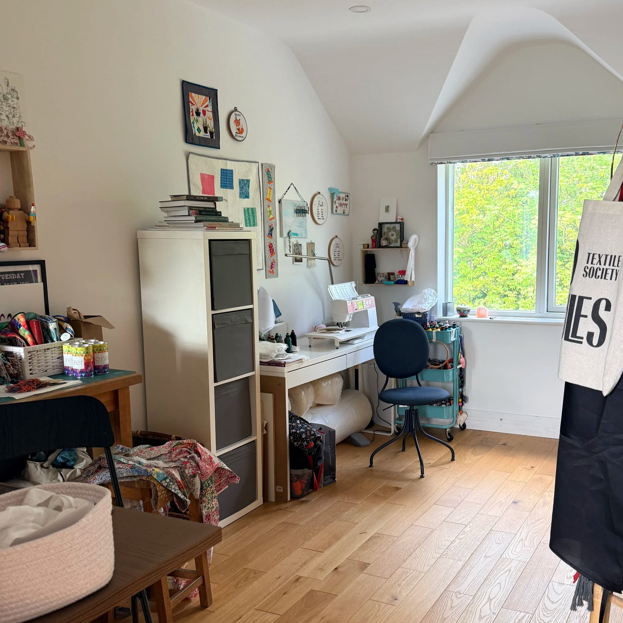 Picture of Anna Hutchinson's home studio. Sewing machine on a desk, with shelving on walls around the room. Textile art on the walls to bring in colour. A working space.