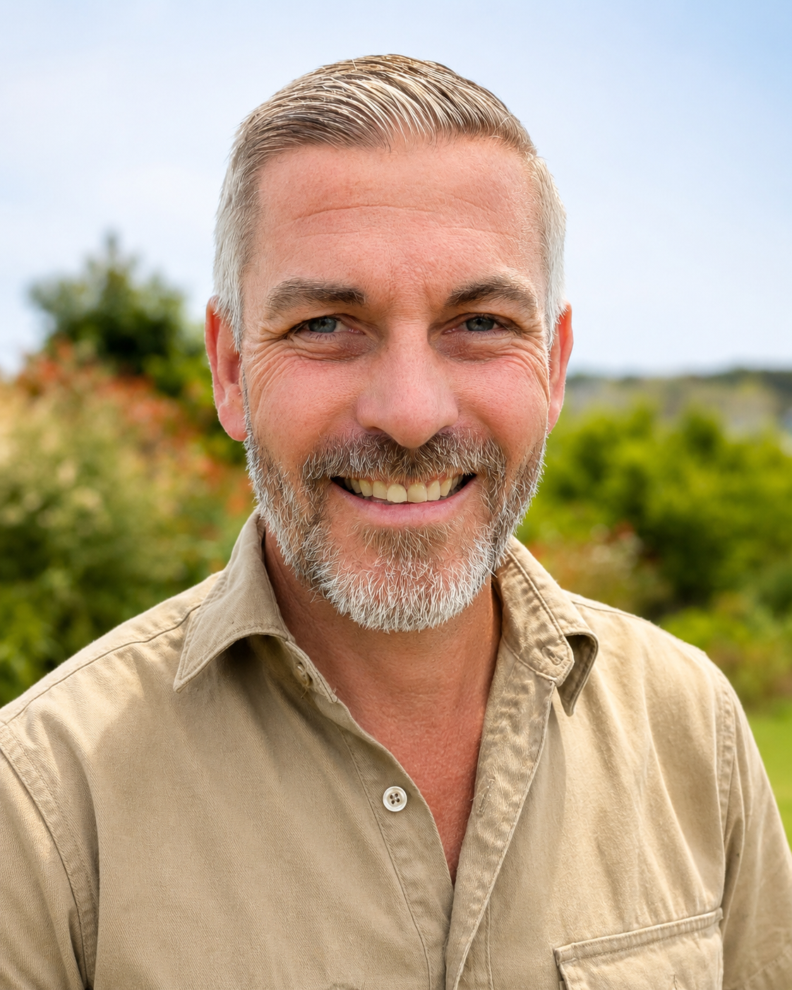 Close-up of a smiling middle-aged man with gray hair and beard, standing outdoors with trees and greenery in the background.