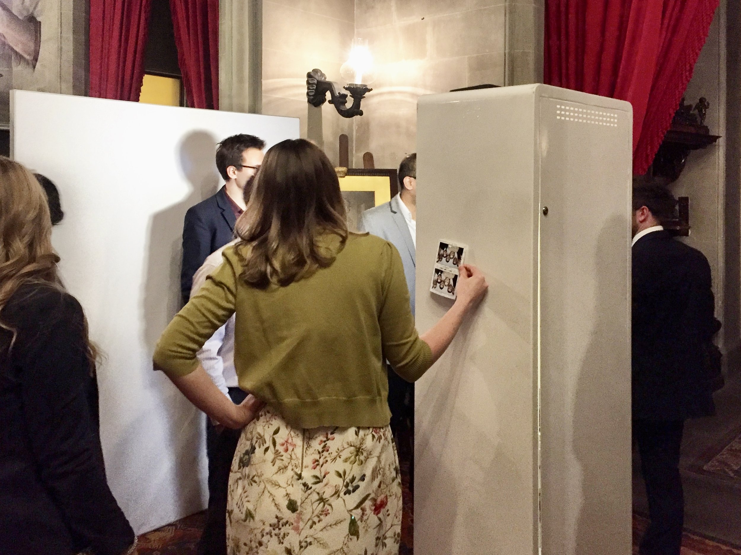 Wedding guests enjoying It’s Social Photo Booth with white backdrop in venue with stone walls and red curtains.