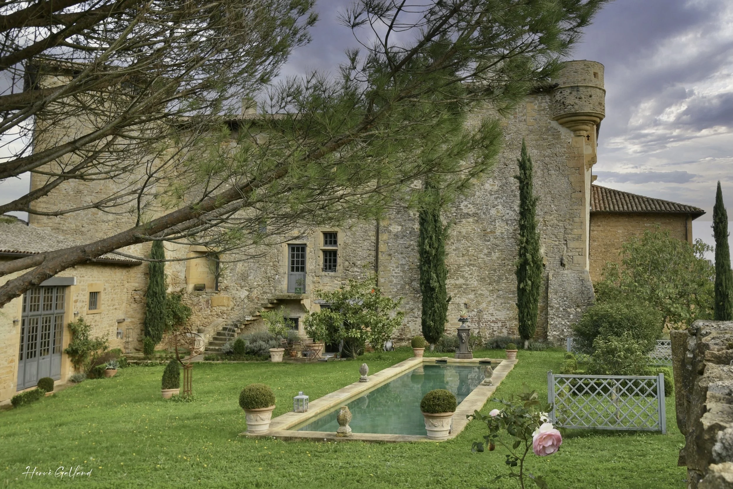 Jardin avec piscine, pelouse verte, arbres, buissons, vieux bâtiment en pierre, fenêtres en verre, escalier, statues, et une rose